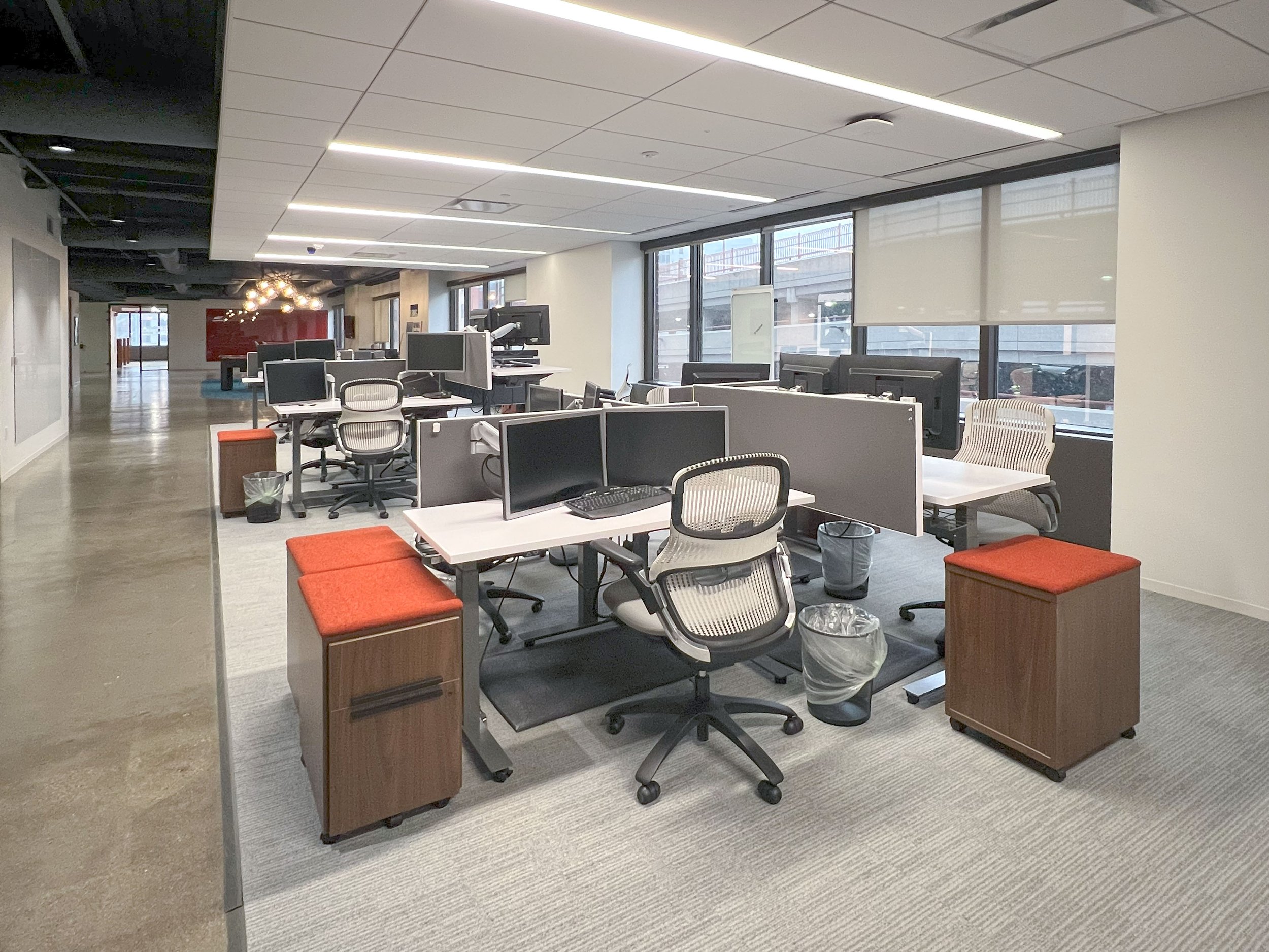 An empty modern office space with multiple desks and chairs, computer monitors, large windows with blinds, and contemporary lighting.