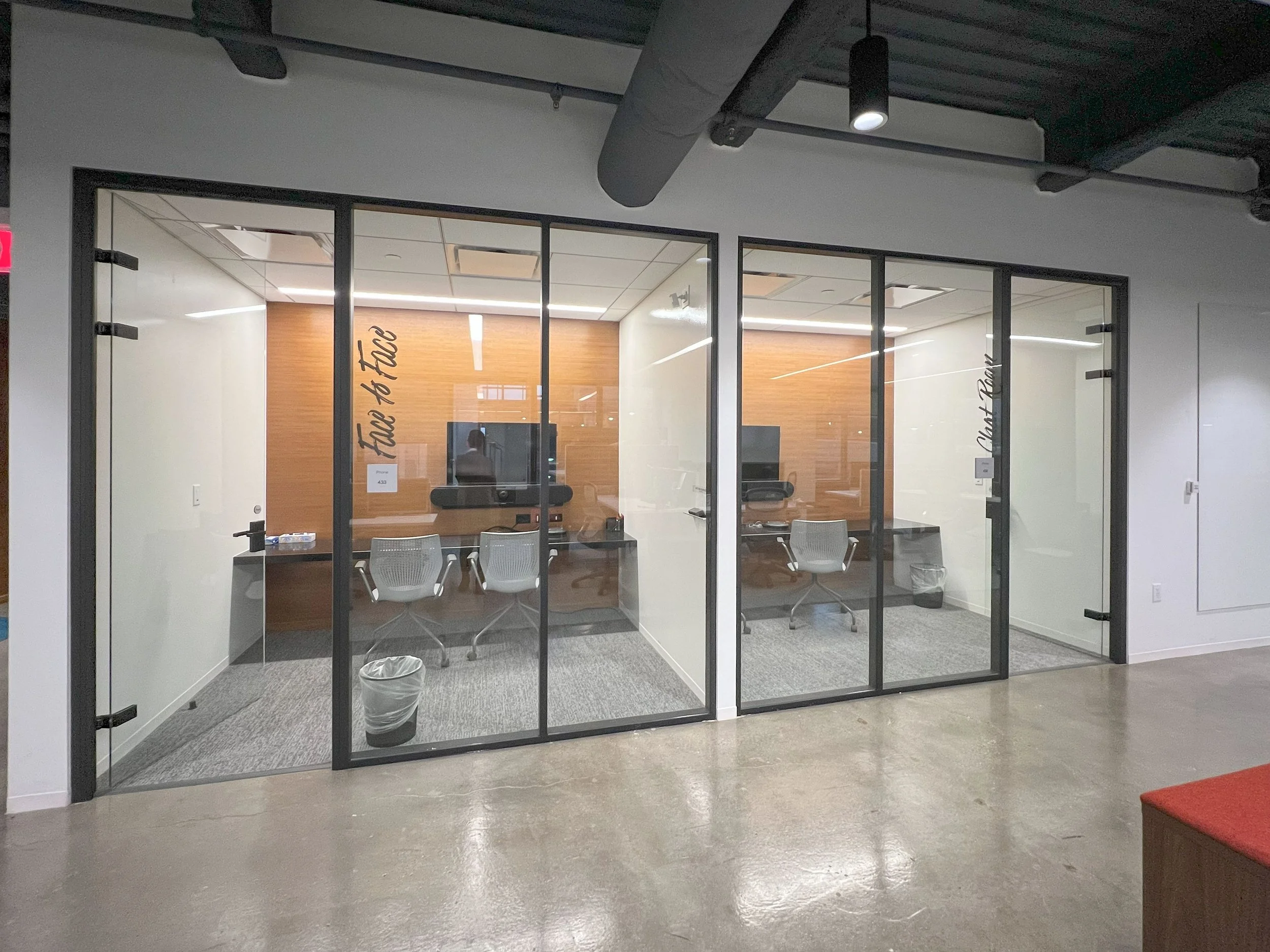 A glass conference room with three chairs, a table, and a spokeswoman inside, with a modern wooden accent wall and ceiling lights.