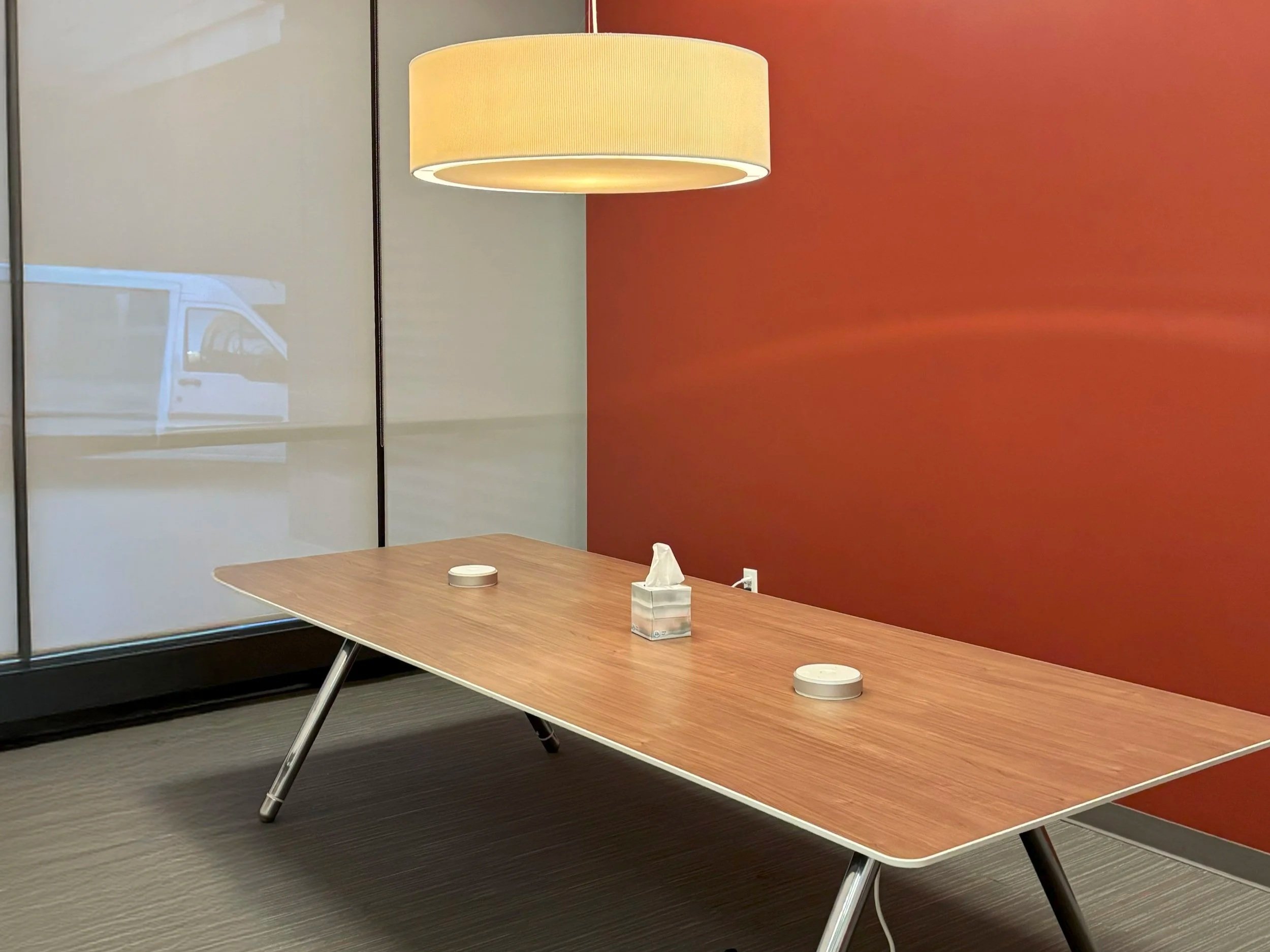 Conference room with a wooden table, tissue box, round coasters, red and white walls, large window, and a hanging ceiling light.