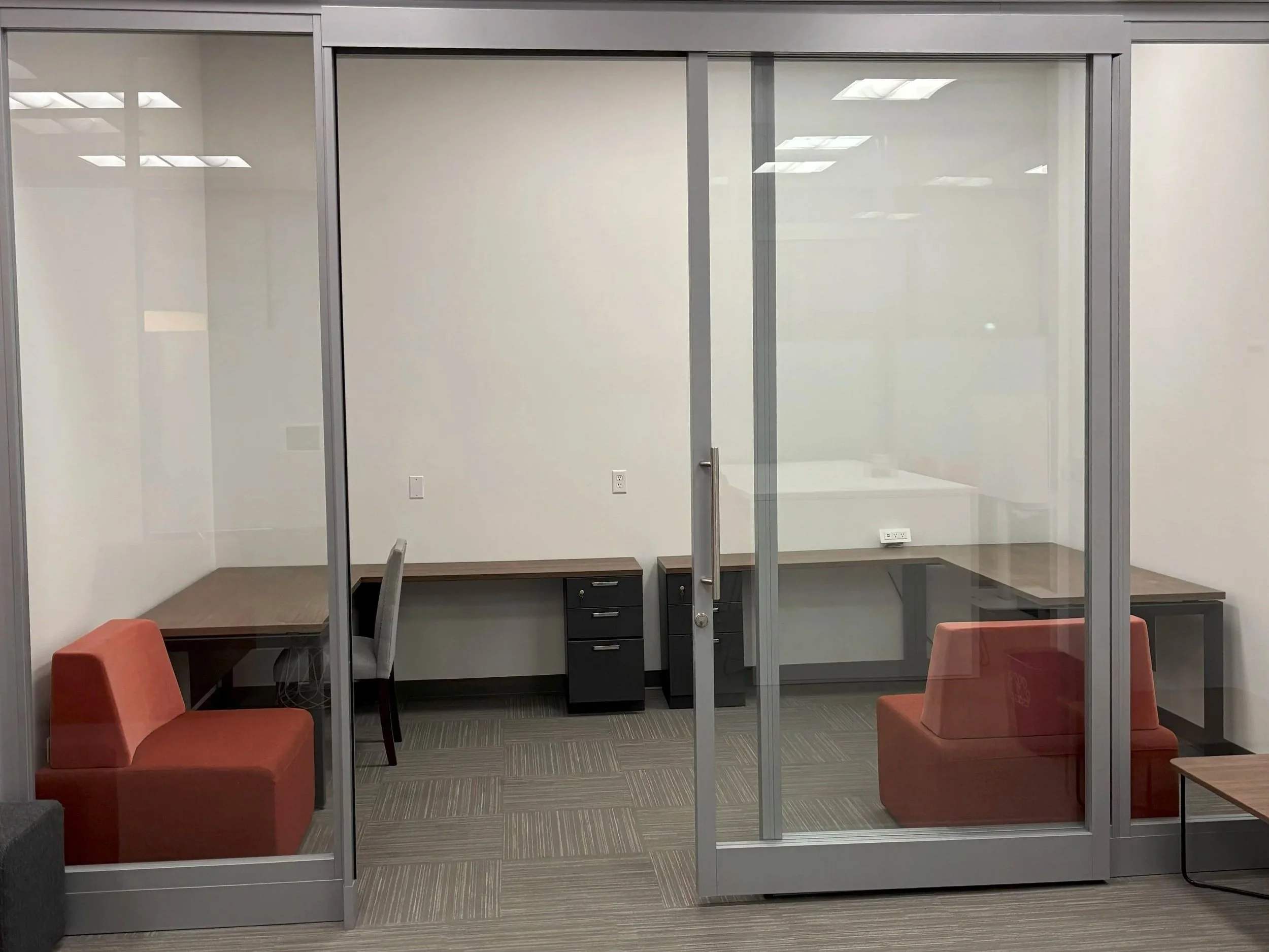 Office cubicle workspace with glass walls, two orange chairs, a desk with a filing cabinet, and a small side table.