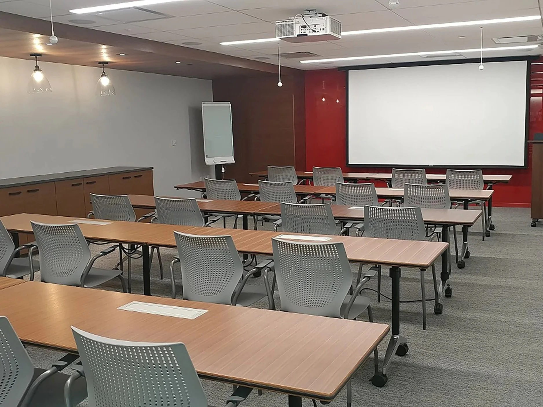 An empty modern conference room with several rows of desks and chairs, a large projection screen on the front wall, ceiling lights, and a flip chart.