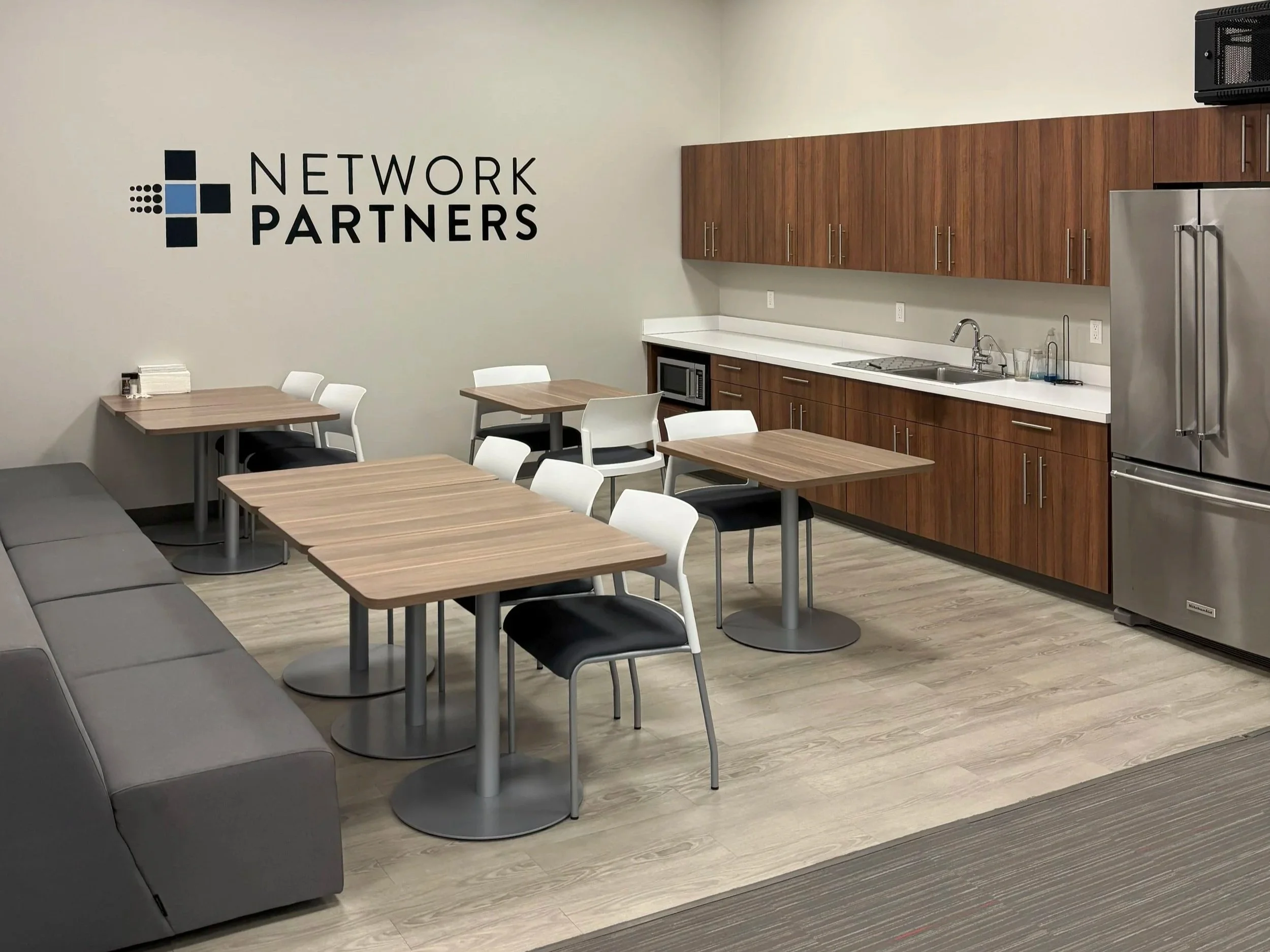 A modern break room with tables and chairs, wooden cabinets, a stainless steel refrigerator, and a wall sign that reads 'Network Partners'.