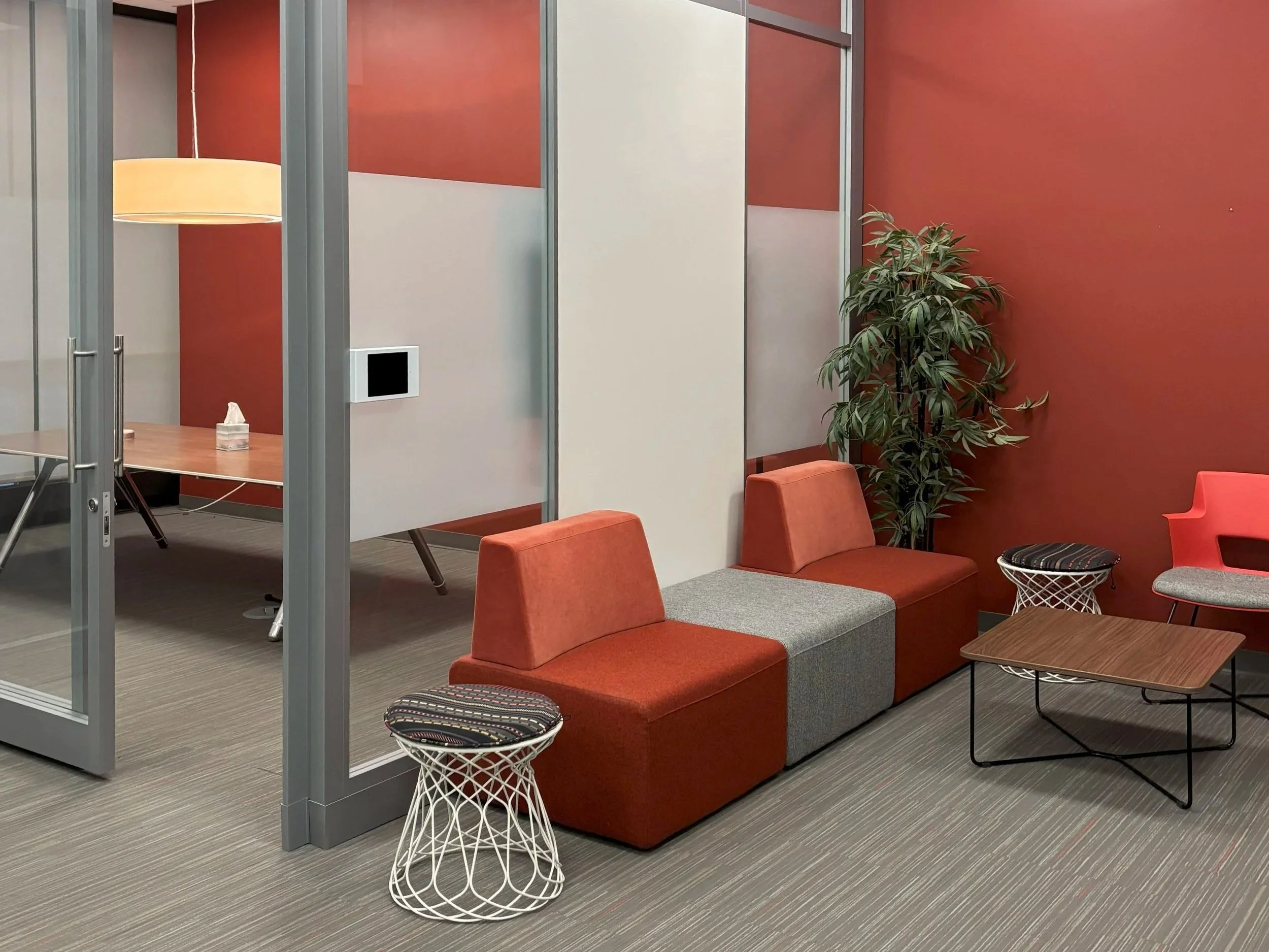 An office lounge area with orange and gray seating, small wireframe side tables, a large potted plant, and cubicle partitions with red and neutral-colored walls.