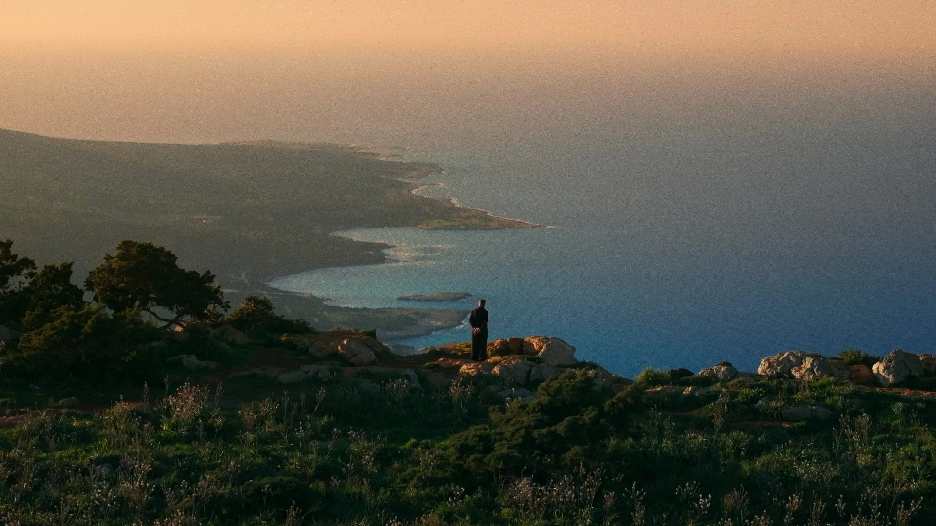 Person standing on a rocky hilltop overlooking a coastal landscape with cliffs, trees, and ocean during sunset.