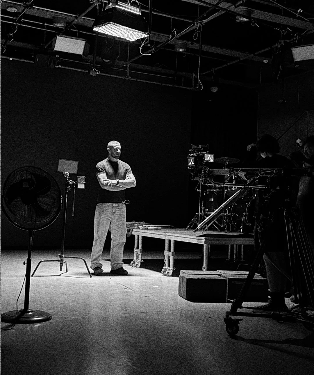 A person with folded arms standing on a stage set, surrounded by film equipment including a camera, lighting, and a large fan, in a studio or theater environment.