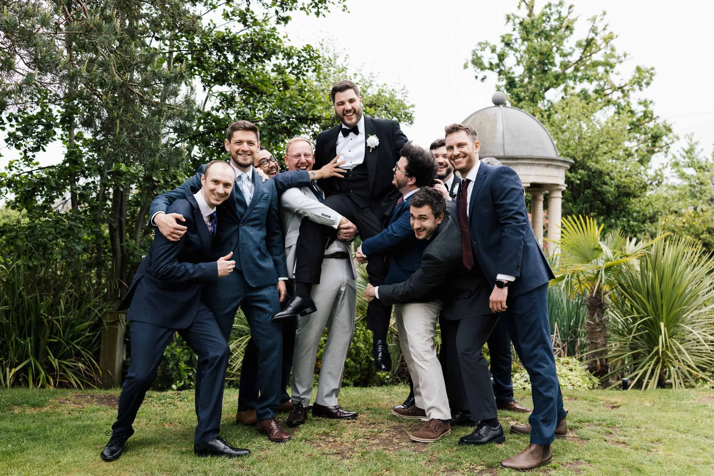 A group of men, dressed in suits and tuxedos, surrounding a man in a tuxedo with a white flower boutonniere, lifting him up in celebration outdoors. They are smiling and appear joyful, with greenery and a gazebo in the background.