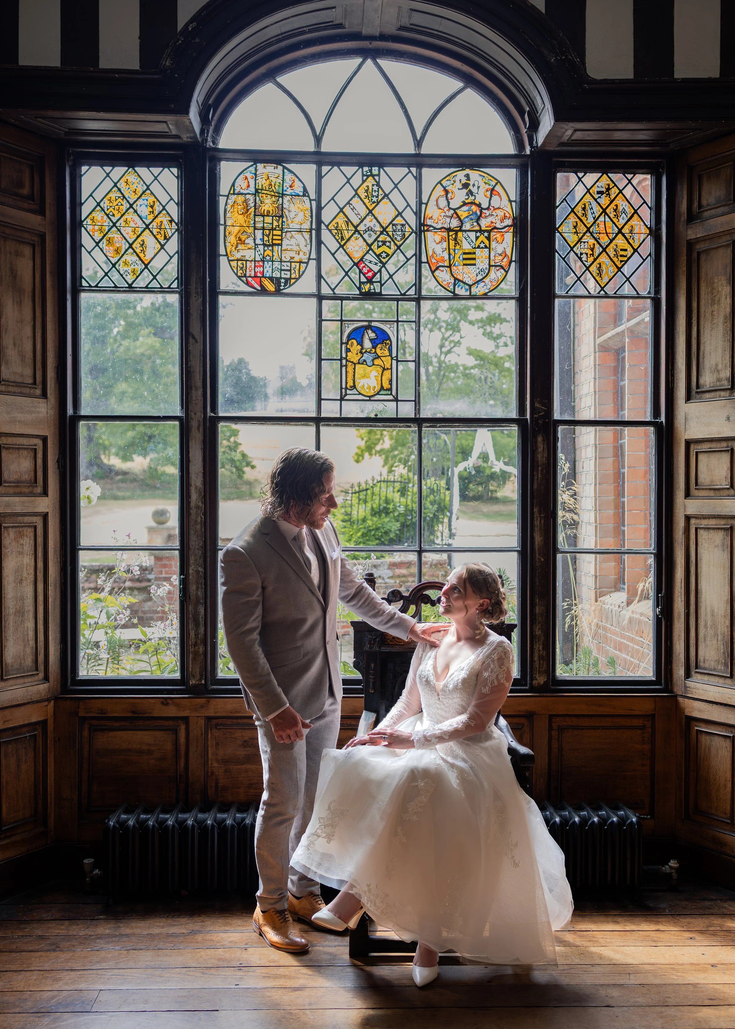 A man and woman in wedding attire inside a room with large stained glass window featuring heraldic shields, with the woman sitting on a chair and the man standing beside her touching her shoulder.