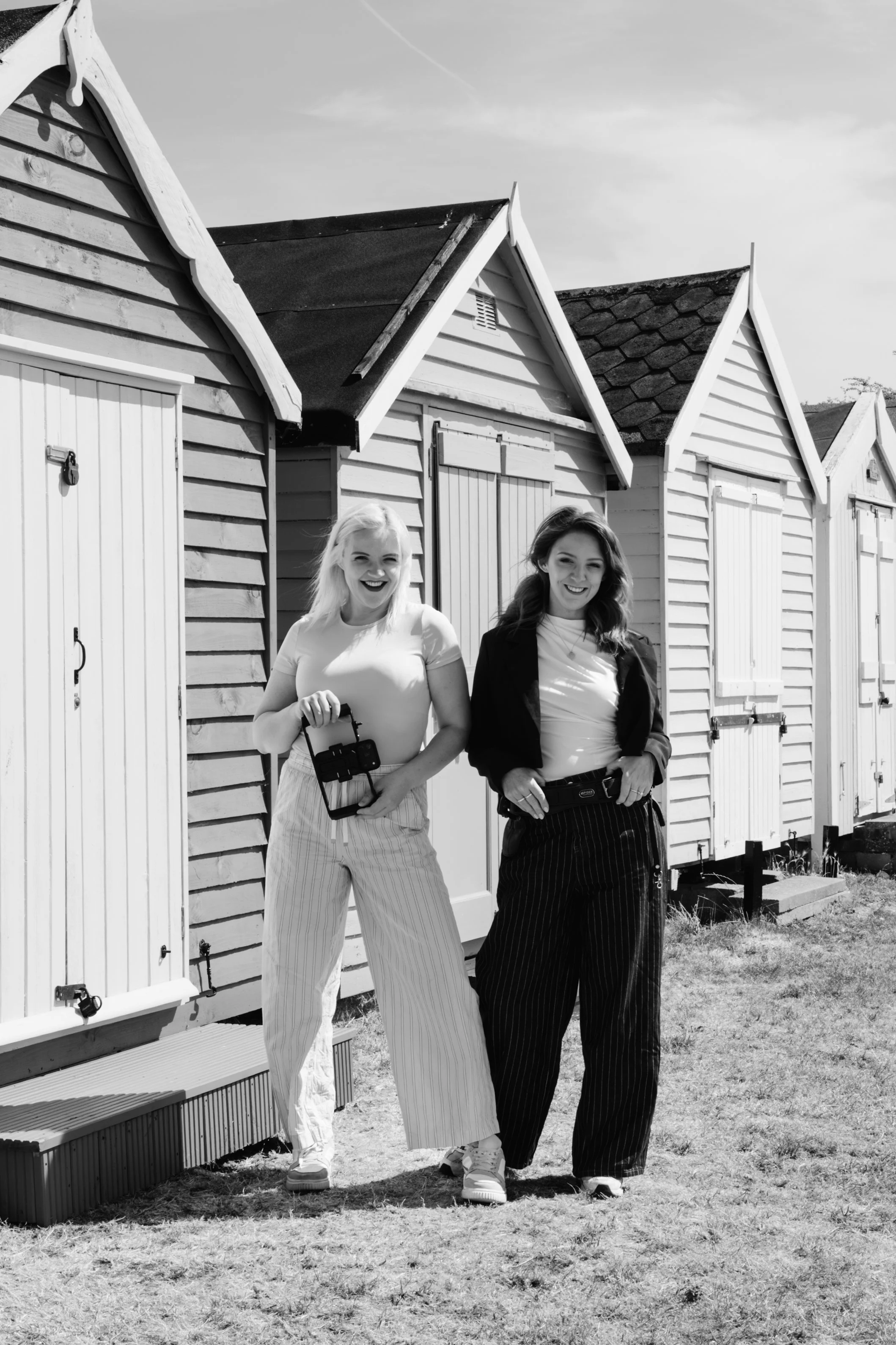 Two women standing outdoors in front of small wooden beach huts, smiling and holding cameras, in black and white photo.
