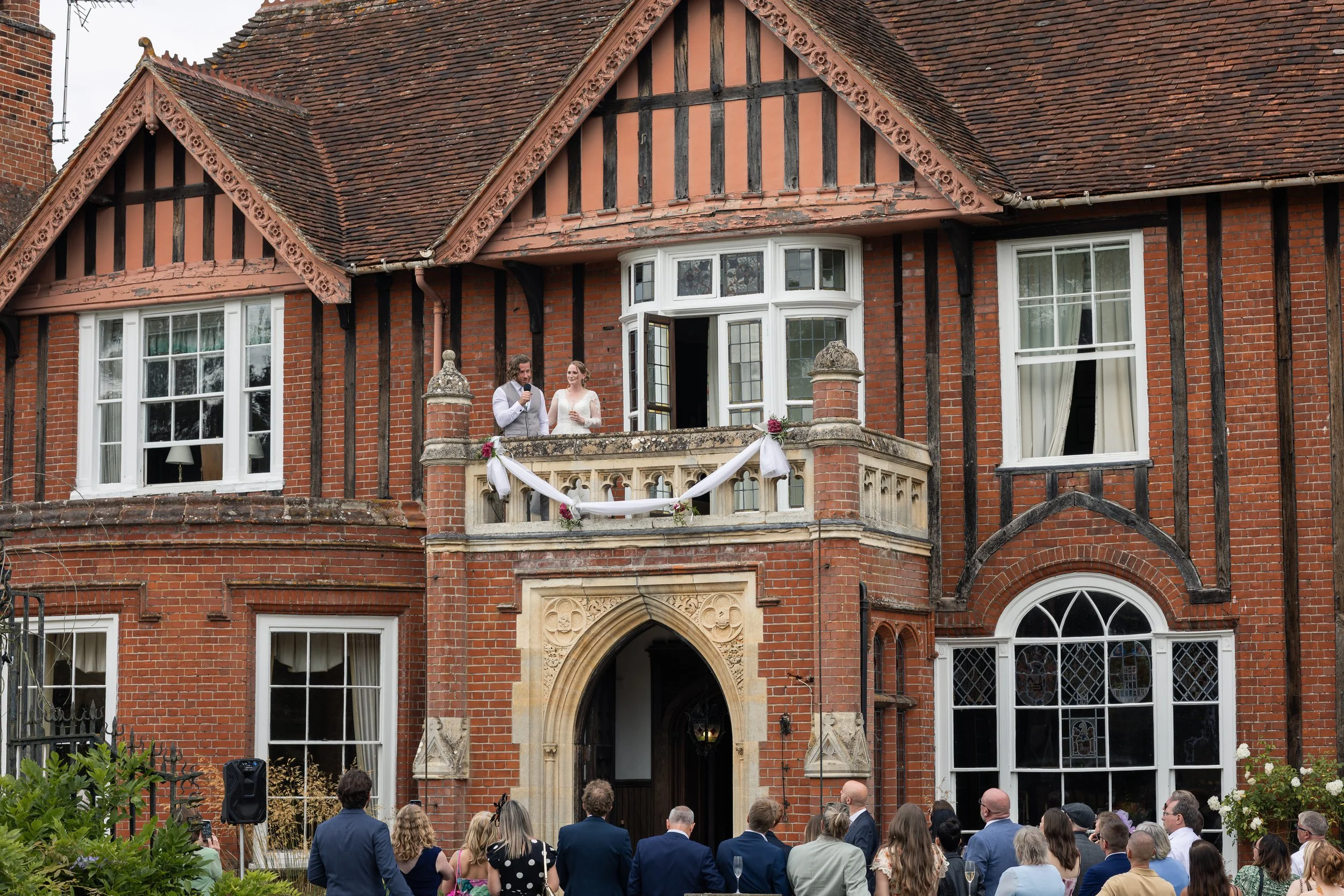 A wedding ceremony outside a large red brick house with a bay window and a balcony. The bride and groom are standing on the balcony, speaking into microphones, while guests are gathered below watching the ceremony.