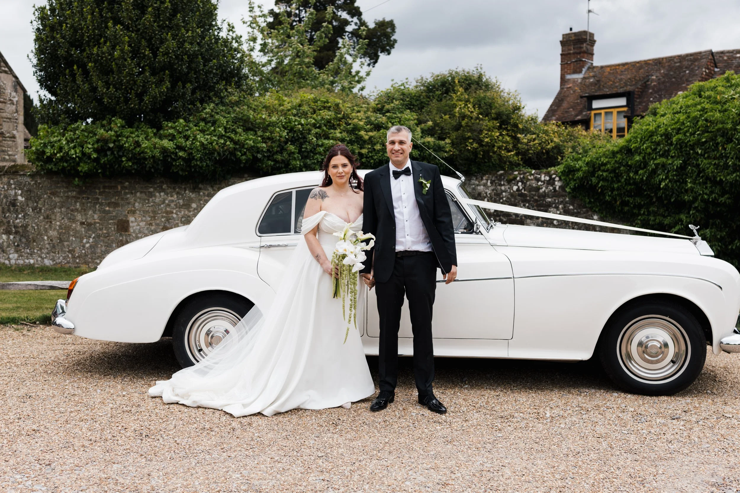 A bride and groom standing in front of a white vintage car on a gravel driveway, with trees and buildings in the background. The bride wears a white wedding gown and holds a bouquet of white flowers, while the groom wears a black tuxedo and bow tie.