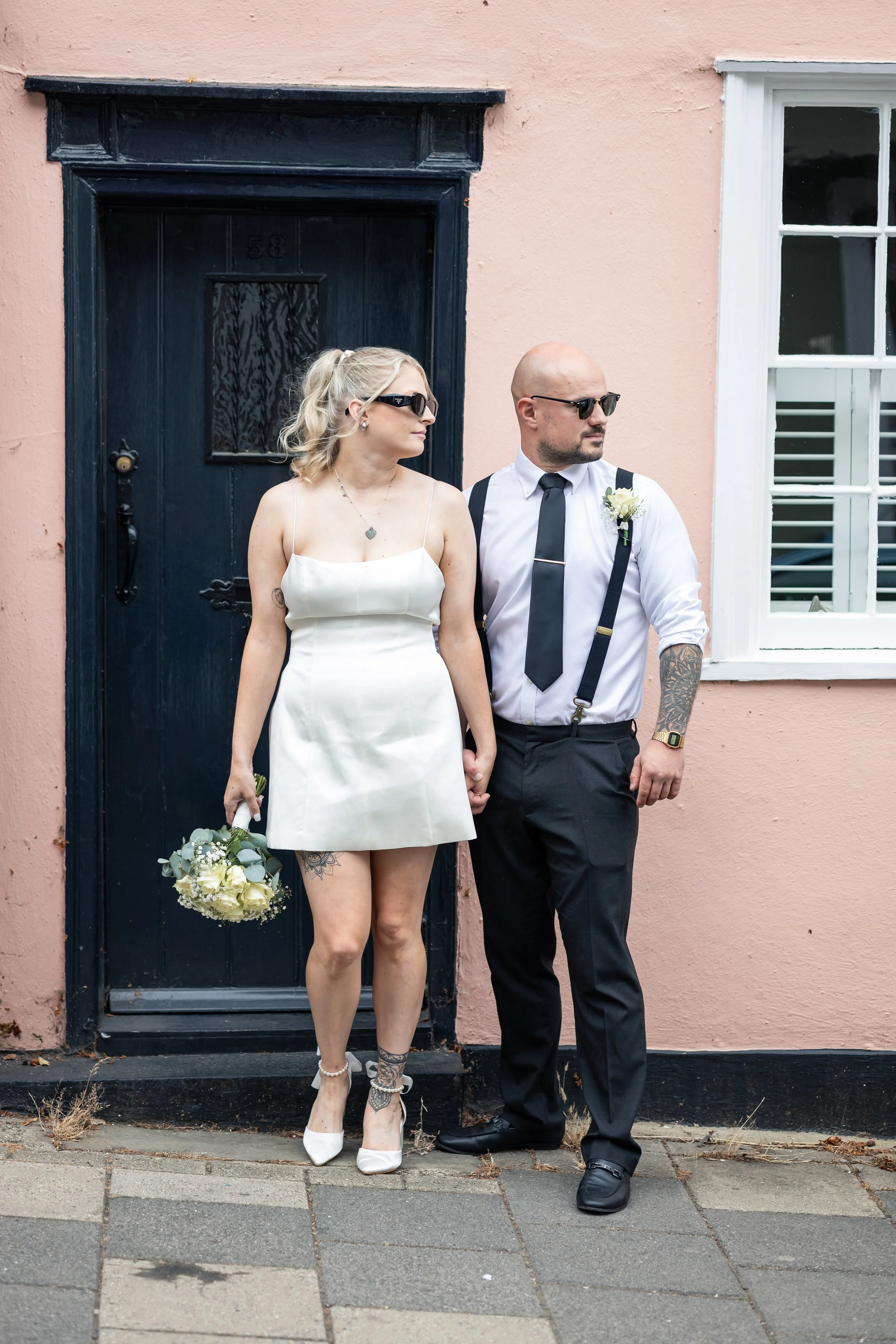 A couple in wedding attire standing on a sidewalk in front of a pink wall with a black door and white window.