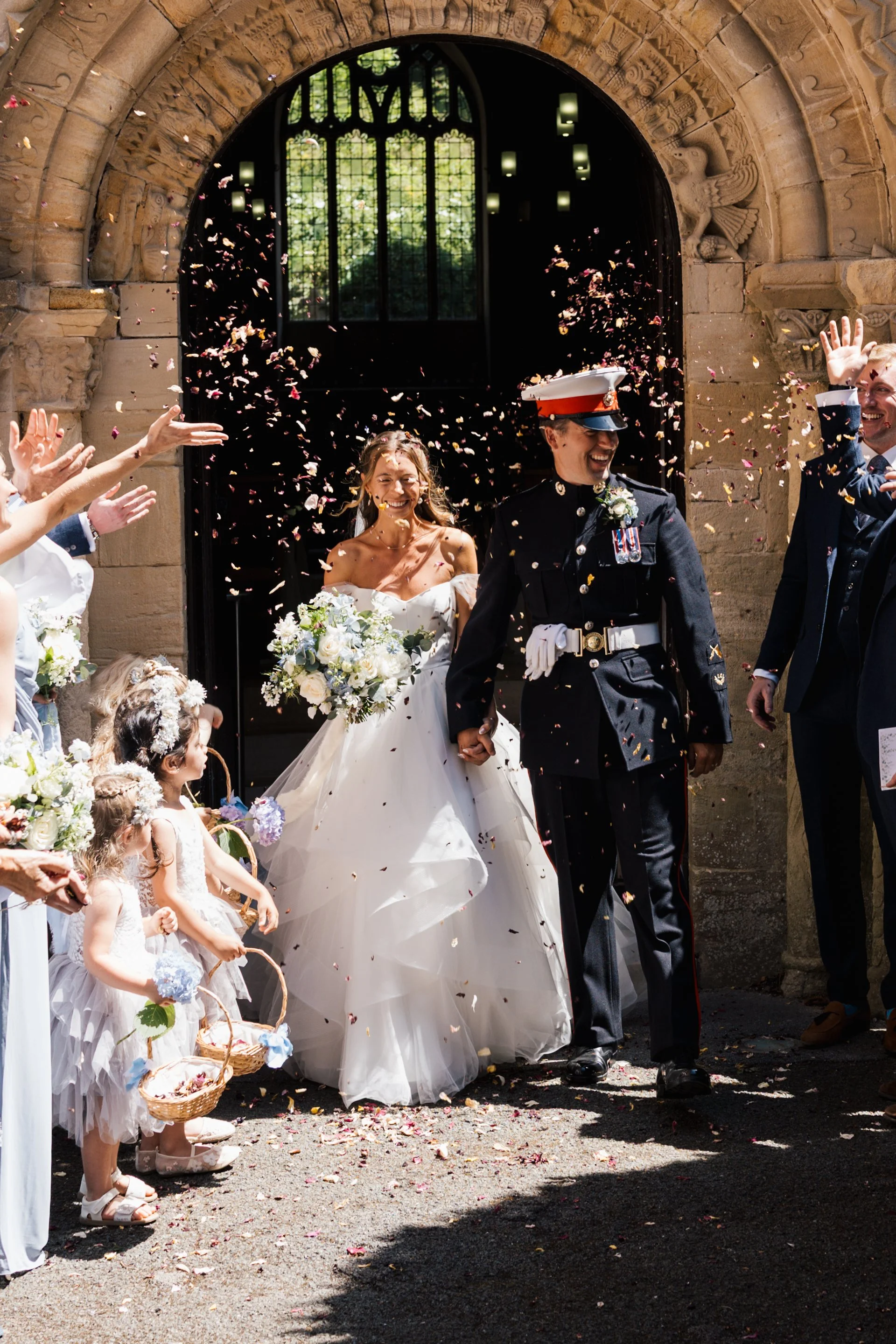 A wedding celebration with a bride in a white gown and a groom in a military uniform walking out of a church, surrounded by flower girls and guests throwing flower petals.