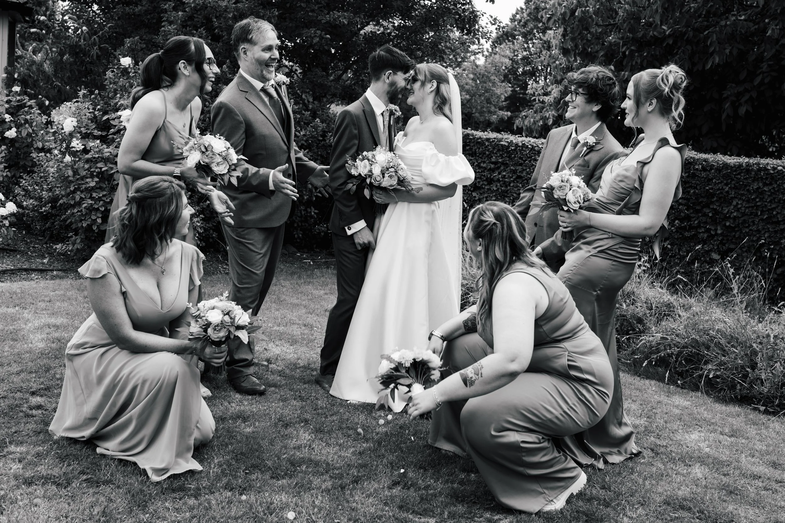 Black and white photo of a wedding ceremony outdoors with guests surrounding the bride and groom. Bride in white dress holding a bouquet, groom in suit with boutonniere. Guests, mostly women in dresses and men in suits, appear joyful, some kneeling, 