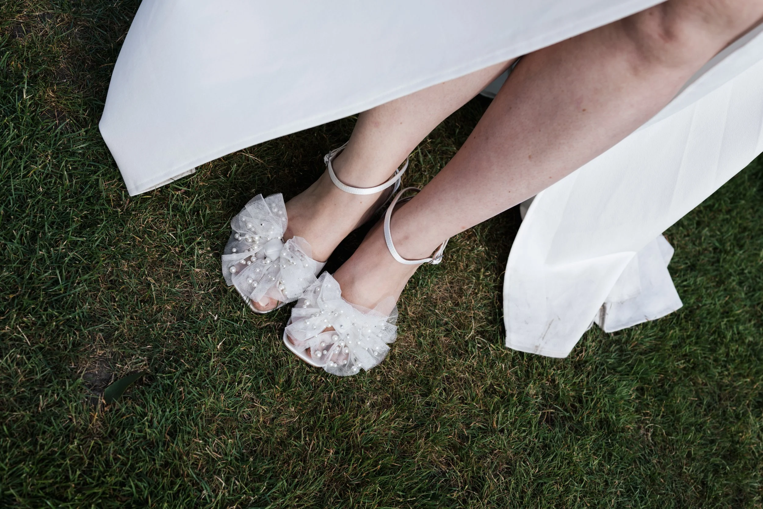 Close-up of a bride's legs and feet, showing her white wedding shoes decorated with pearls and tulle, standing on grass next to a white fabric drape.