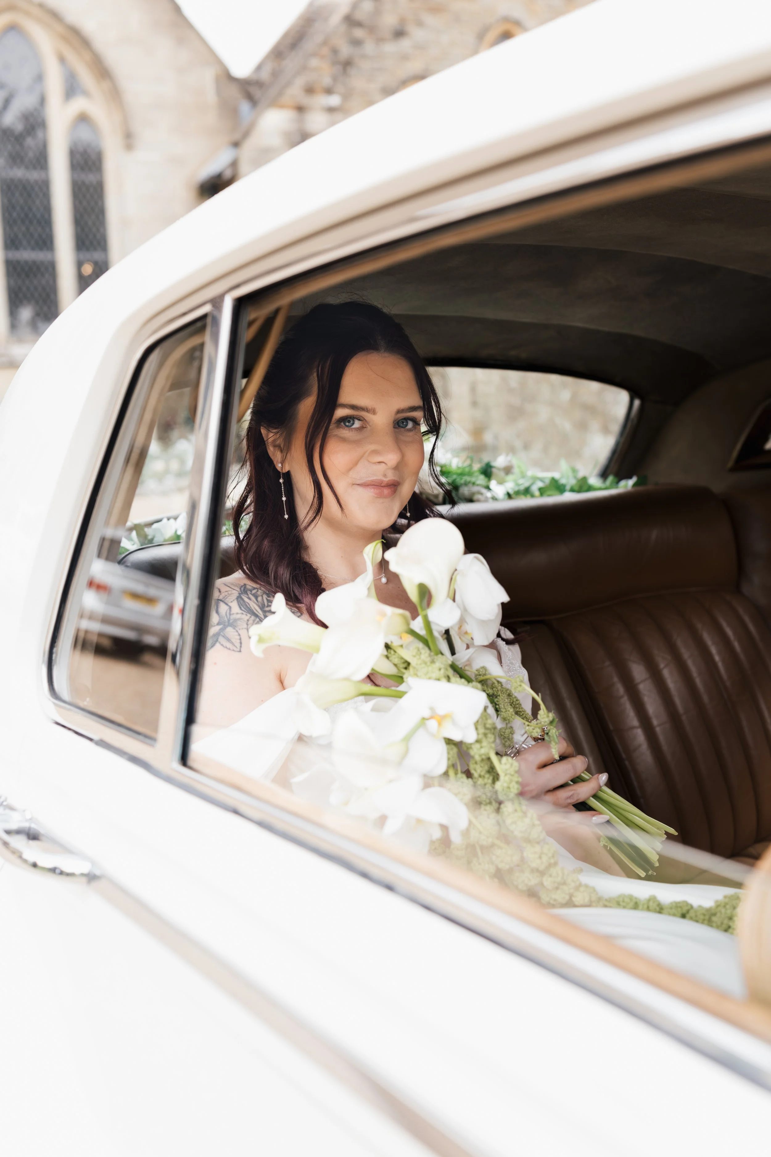 A woman with dark hair and tattoos on her shoulder sitting in the backseat of a vintage white car, holding a bouquet of white flowers, looking out the window.