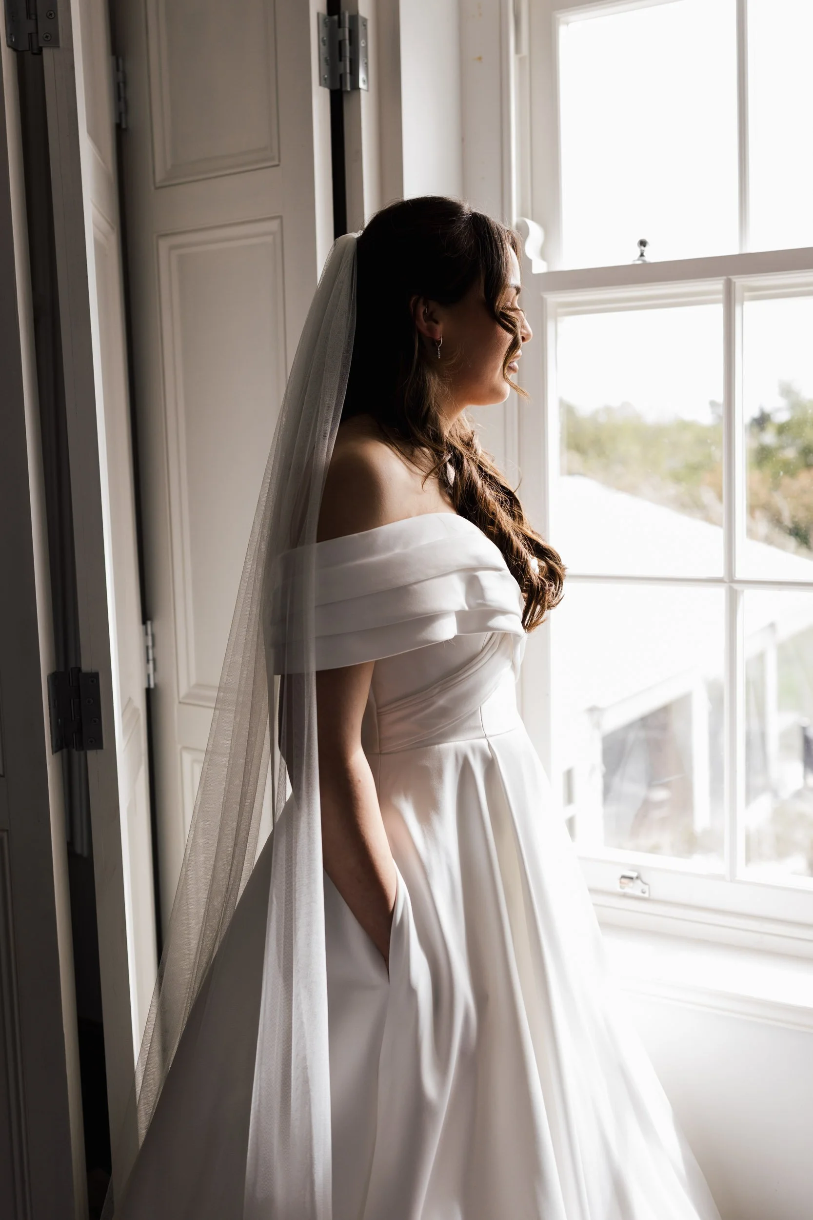 A woman in a white off-shoulder wedding dress with a veil standing by a window, smiling and looking outside, with natural light illuminating her face and dress.