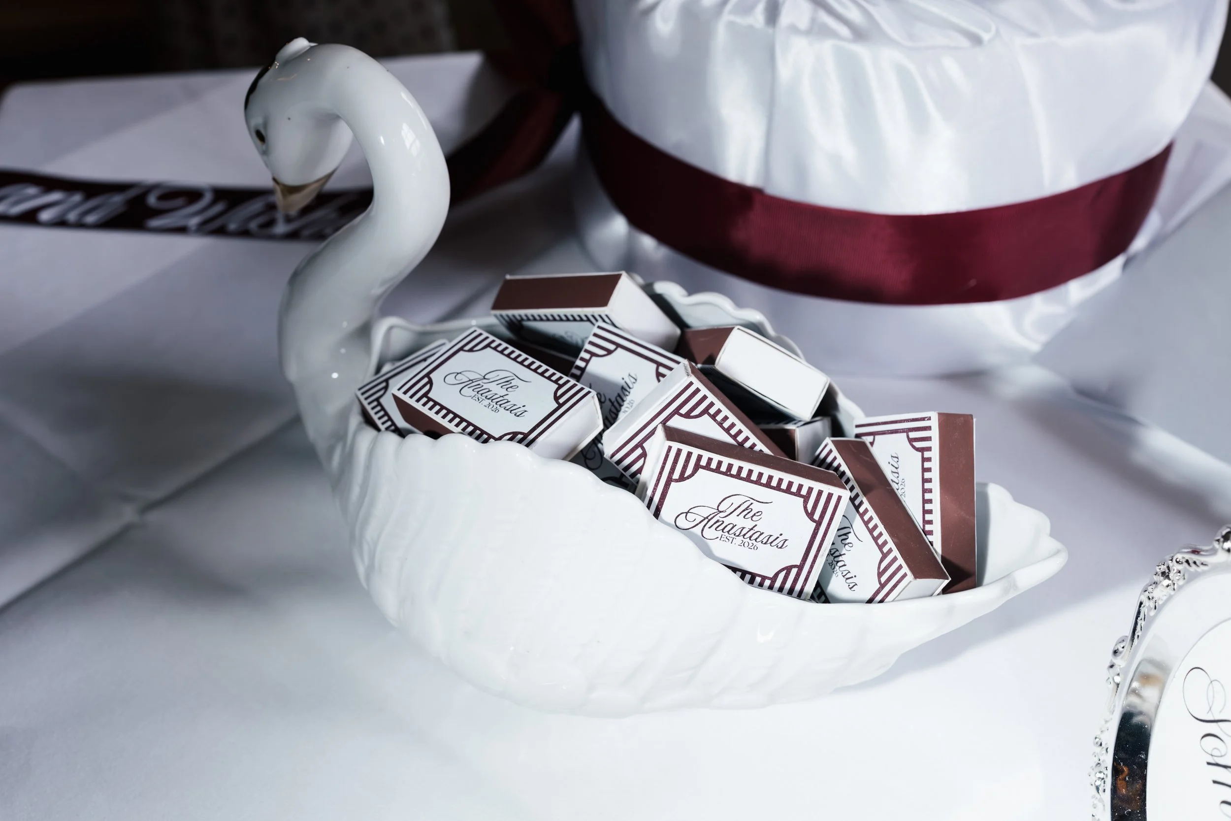 A white ceramic swan-shaped bowl filled with individually wrapped chocolates labeled 'The Anastasias' set on a white table with a white and maroon wrapped gift in the background.