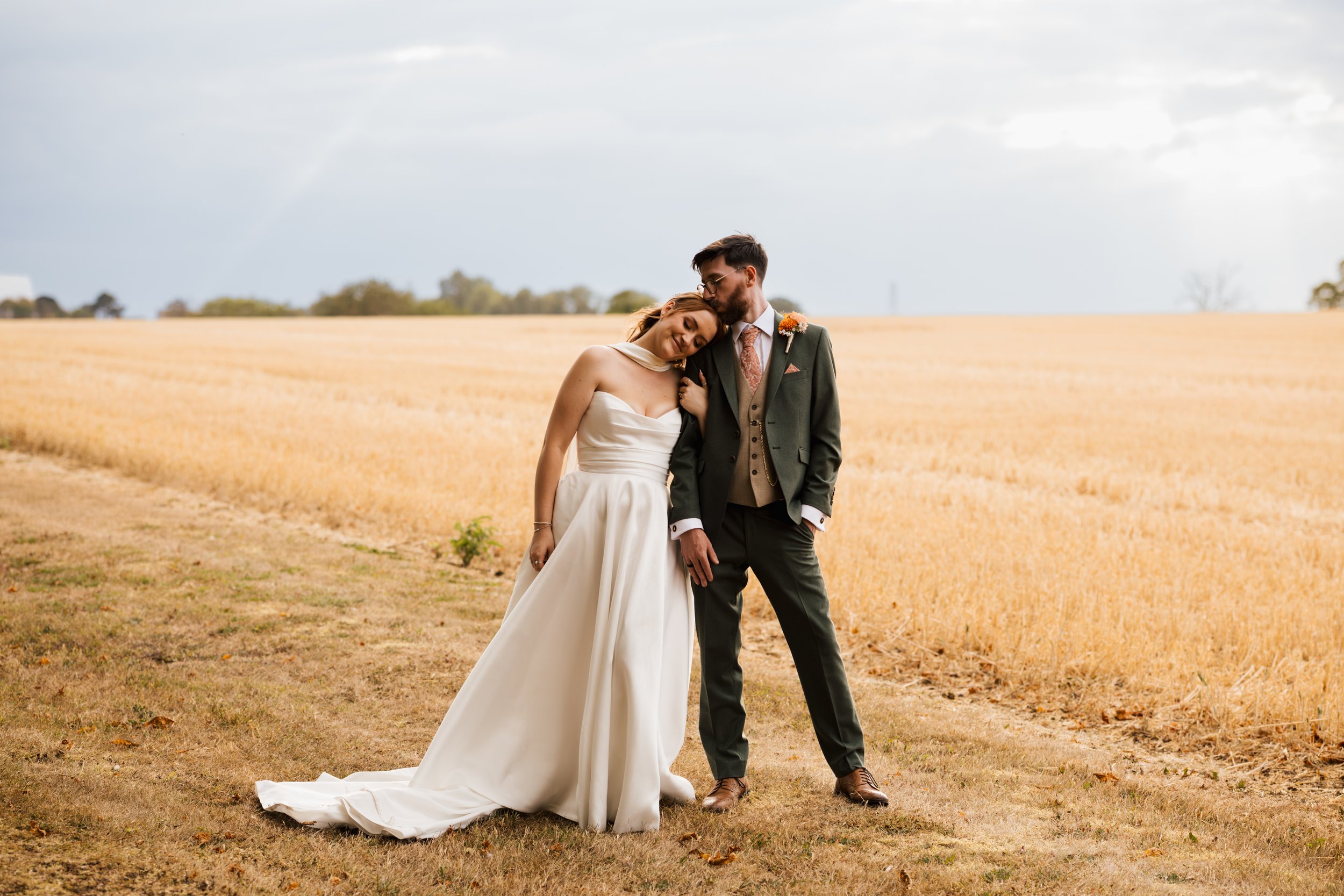 A bride and groom sharing a moment in a golden field under a cloudy sky, with the groom kissing the bride on her head.