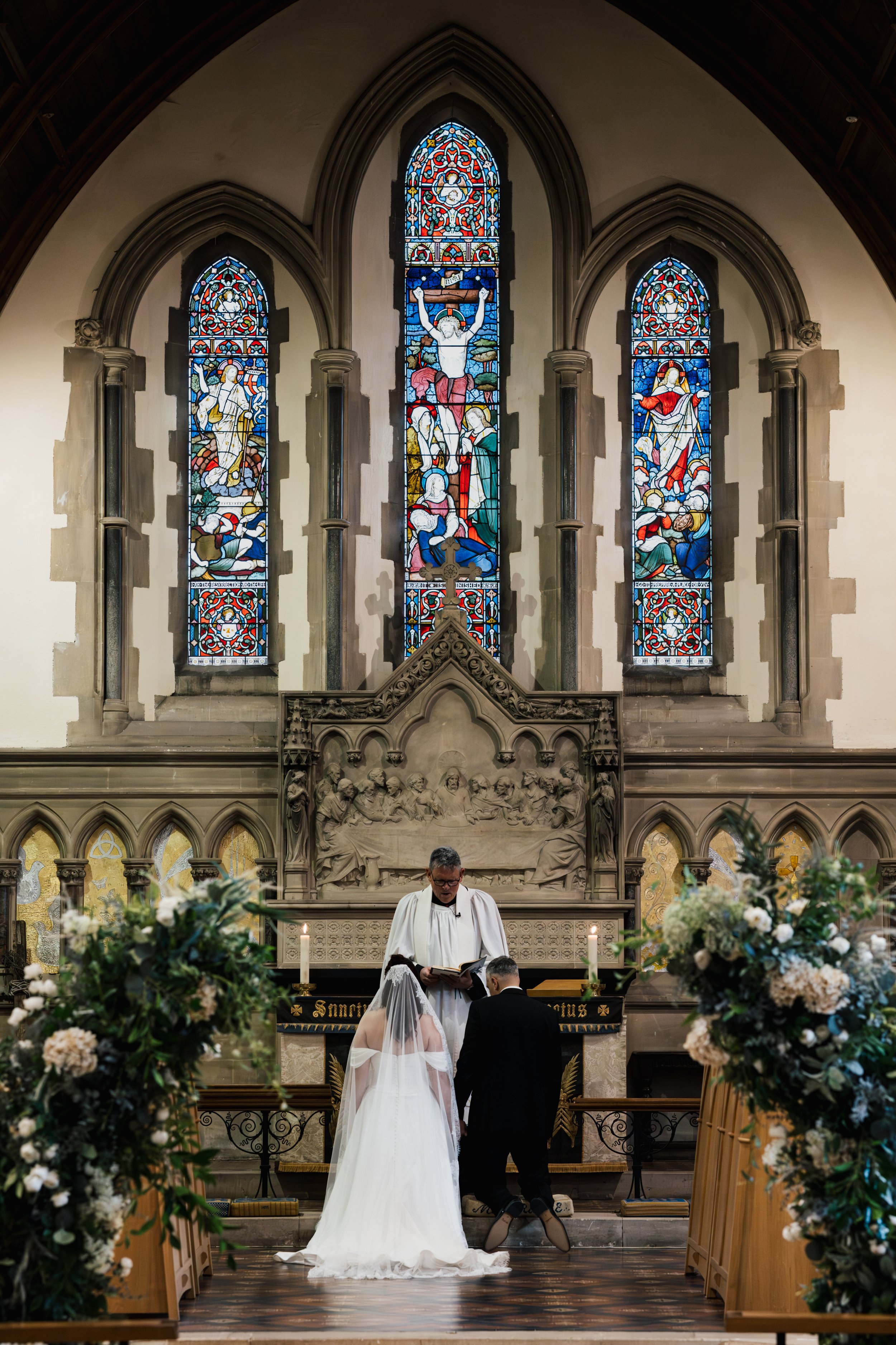 A wedding ceremony taking place inside a church with stained glass windows depicting religious figures. A bride in a white gown and veil and a groom in a black suit stand before a priest during their vows. floral arrangements line the aisle.