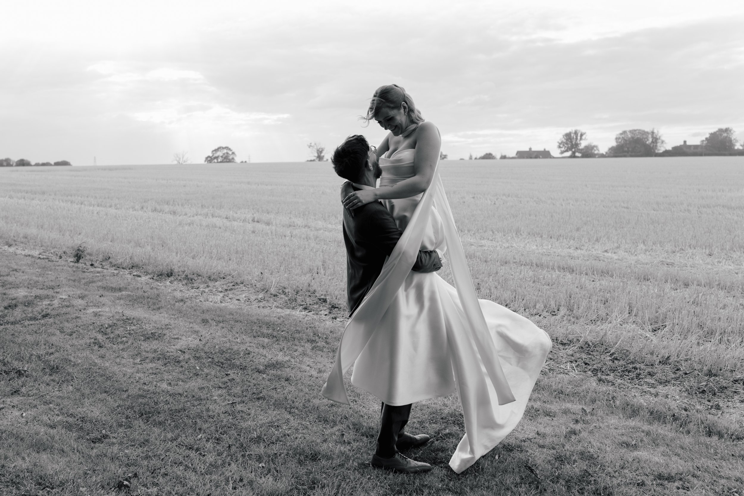 A couple sharing a dance outdoors in an open field, with the woman in a long dress and the man in a suit, both smiling and enjoying the moment.