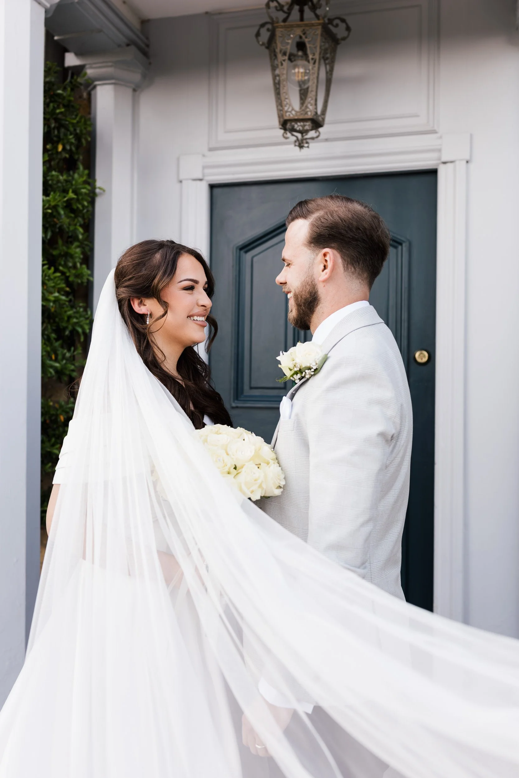 A bride and groom smiling and looking at each other, standing in front of a blue door outside a house, with the bride holding a bouquet of white roses and wearing a veil, wedding attire, and the groom wearing a light-colored suit with a boutonniere.