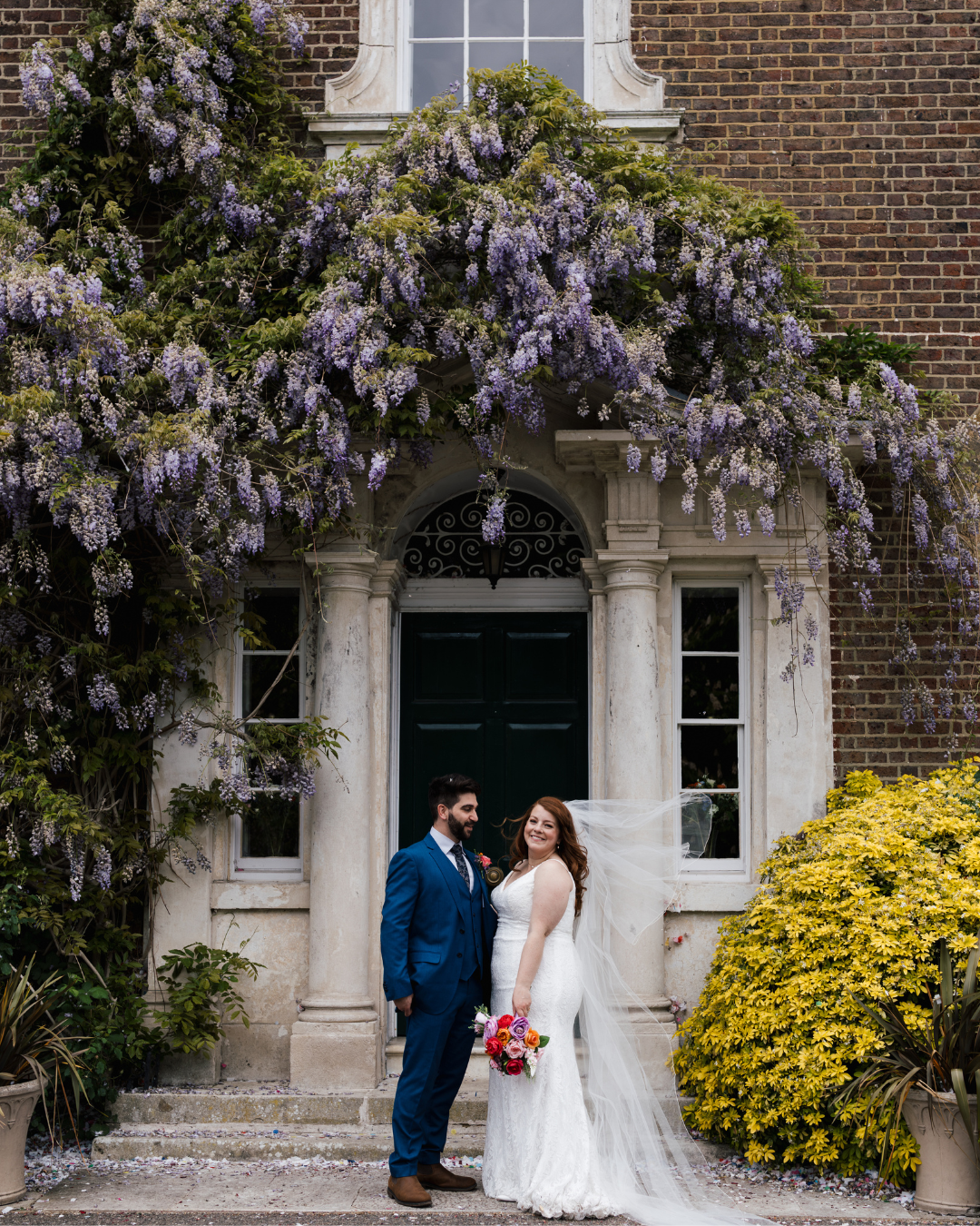 A happy bride and groom standing in front of a historic building with purple flowering vines and colorful bushes.