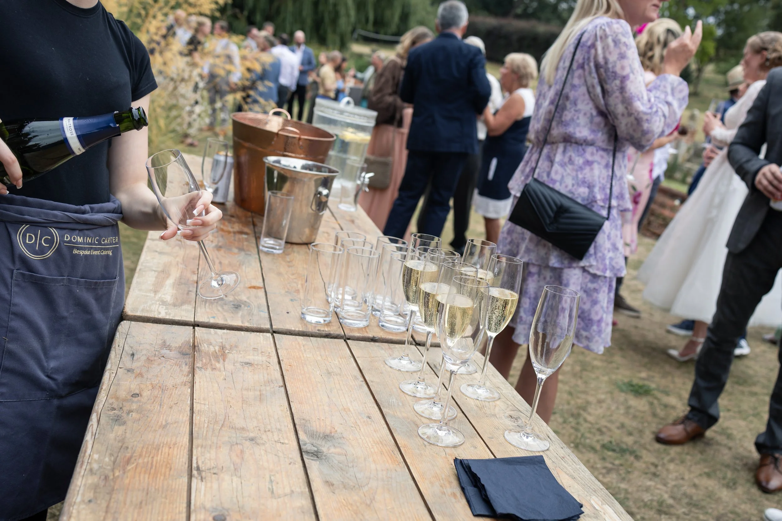 Guests at an outdoor event served champagne from a wooden table with glasses; some people are mingling in the background.
