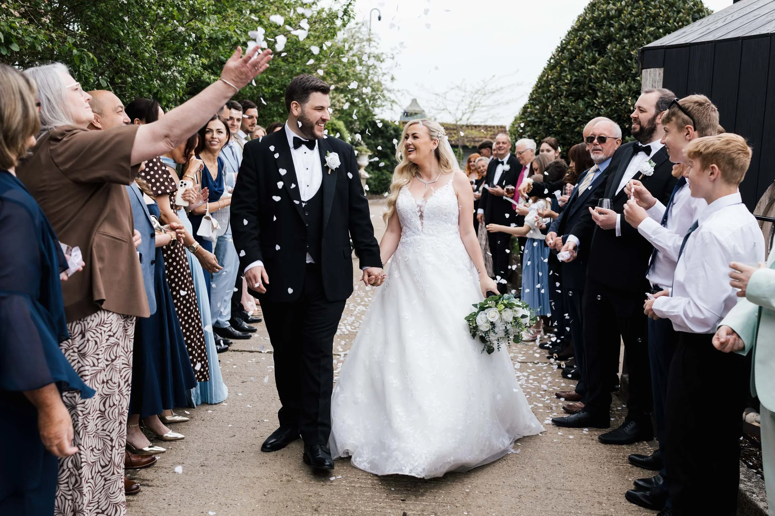 A bride and groom holding hands and smiling at each other during their wedding ceremony, surrounded by friends and family, outdoors with greenery and overcast sky.