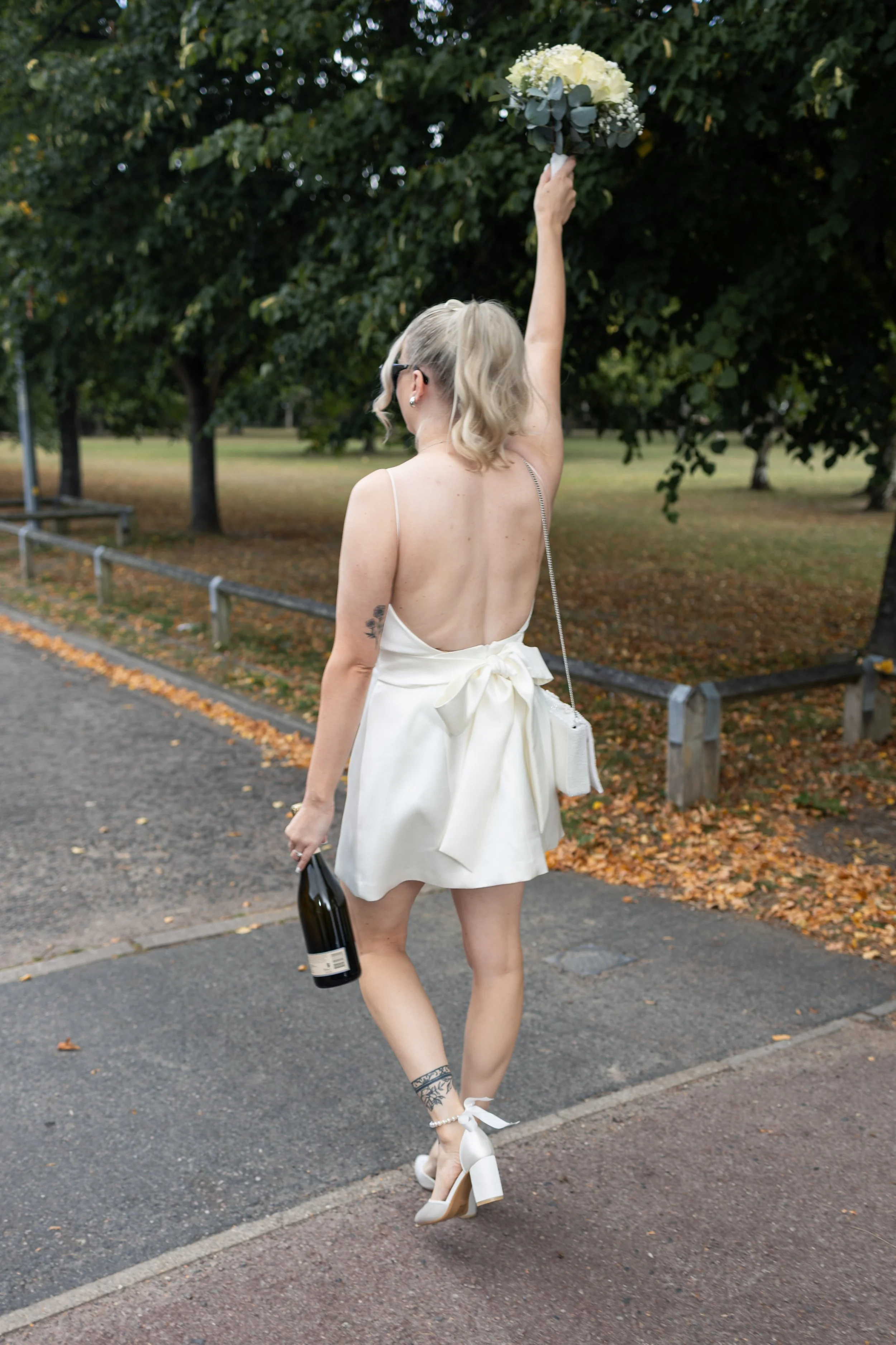 A woman in a backless white dress with a large bow, white high heels, and tattoos, holding a champagne bottle in one hand and a bouquet in the other, walking outdoors on a pathway lined with autumn leaves.
