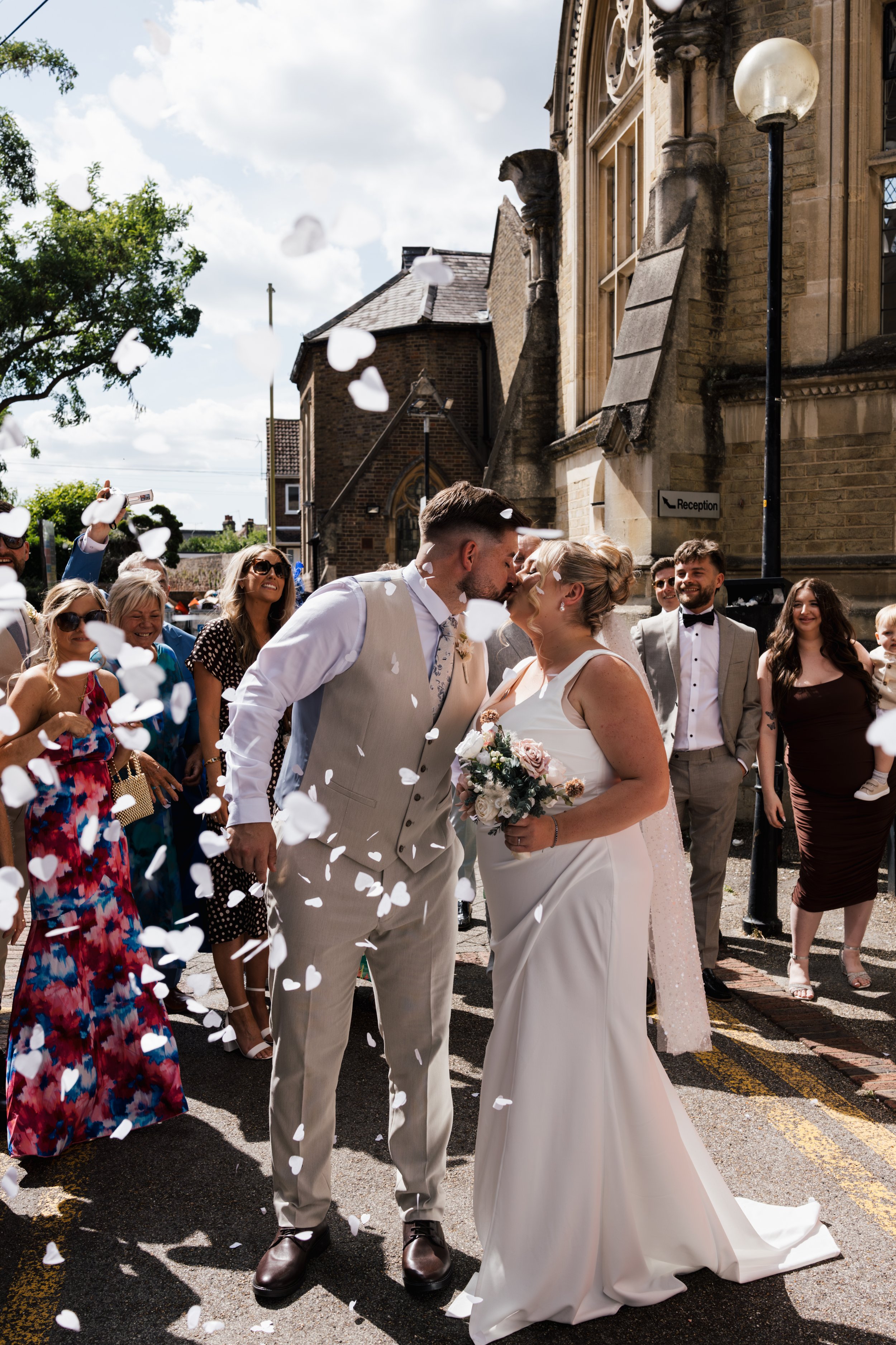 A newlywed couple kiss in front of smiling guests as white paper hearts fall around them outside a church on a sunny day.