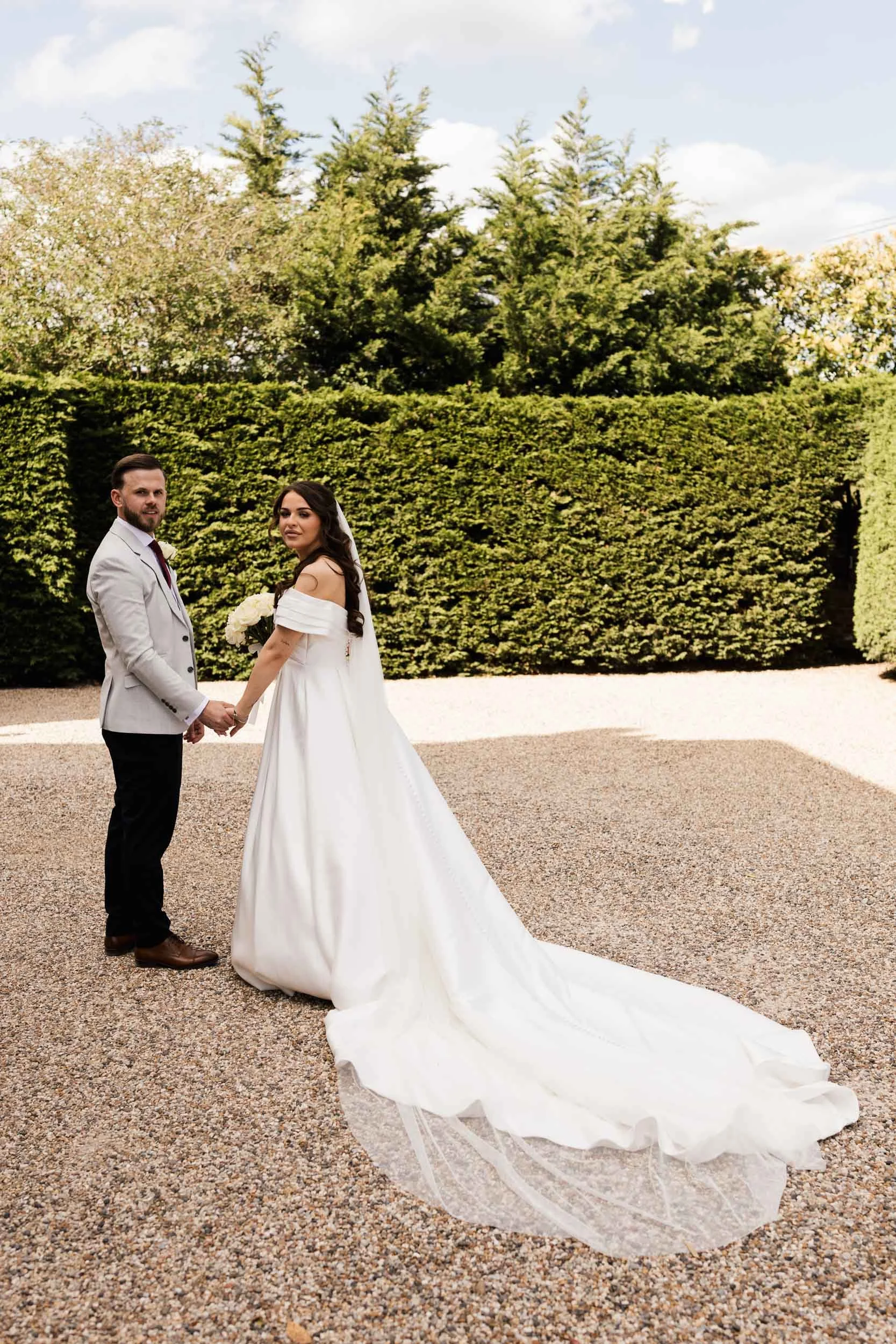 A bride and groom stand hand in hand outdoors on a pathway, with a neatly trimmed hedge and tall trees in the background, during daytime. The bride wears a white off-the-shoulder wedding gown with a long train, and the groom wears a light gray suit w