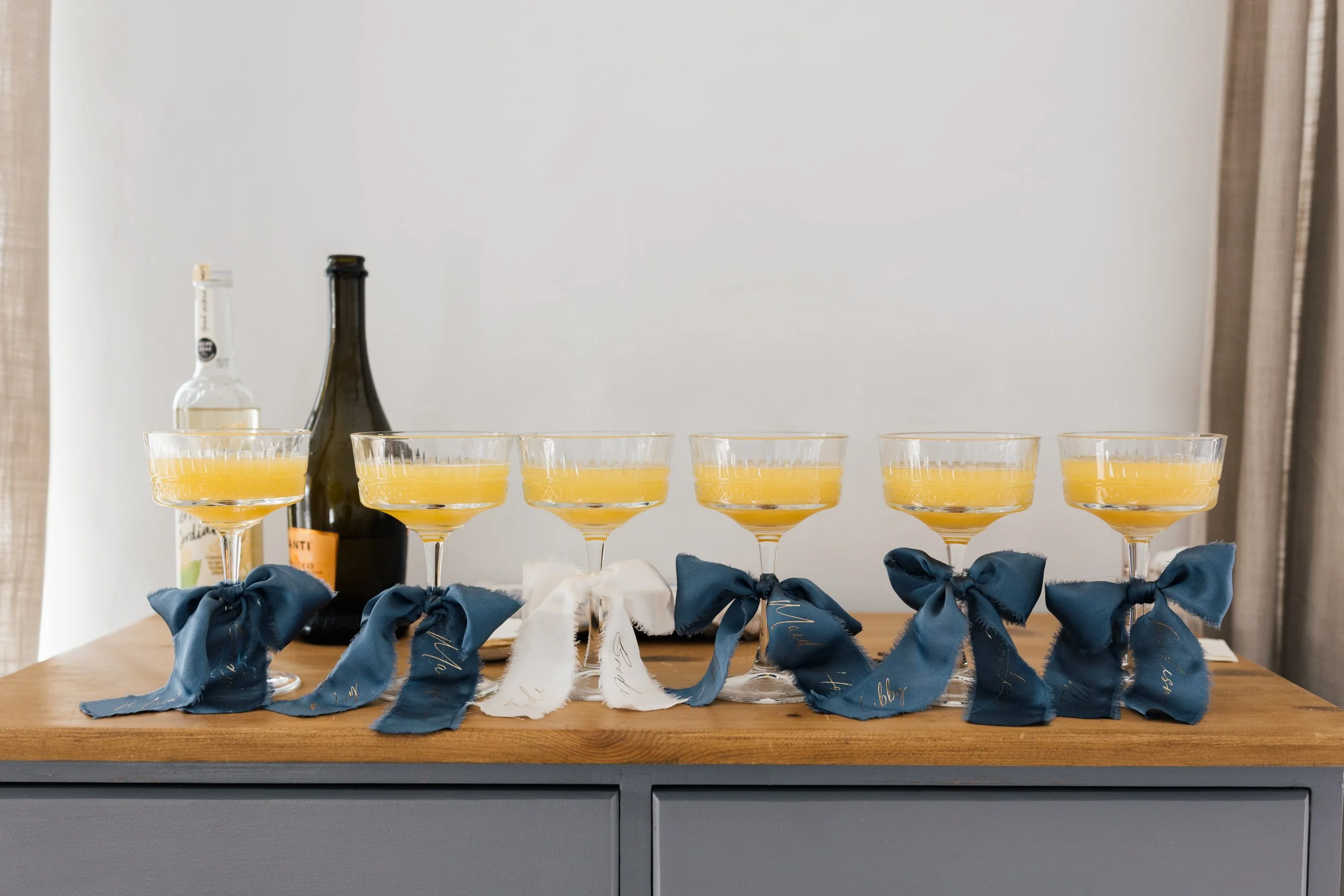 Six champagne glasses filled with a yellow beverage, decorated with navy blue bows, placed on a wooden surface with substance tags hanging from the bows. Two bottles of alcohol are in the background, one clear and one dark-colored, on a plain white w