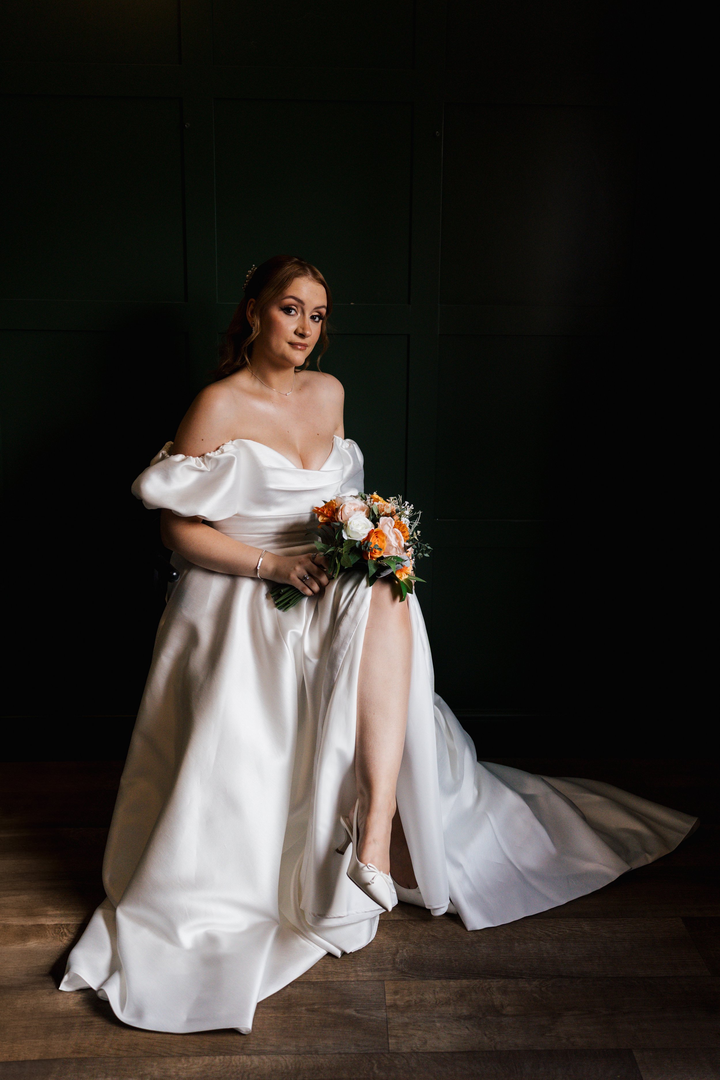 A woman in a white wedding dress sitting on a wooden floor holding a bouquet of orange and white flowers, with her right leg crossed over her left and her right foot in a white shoe.
