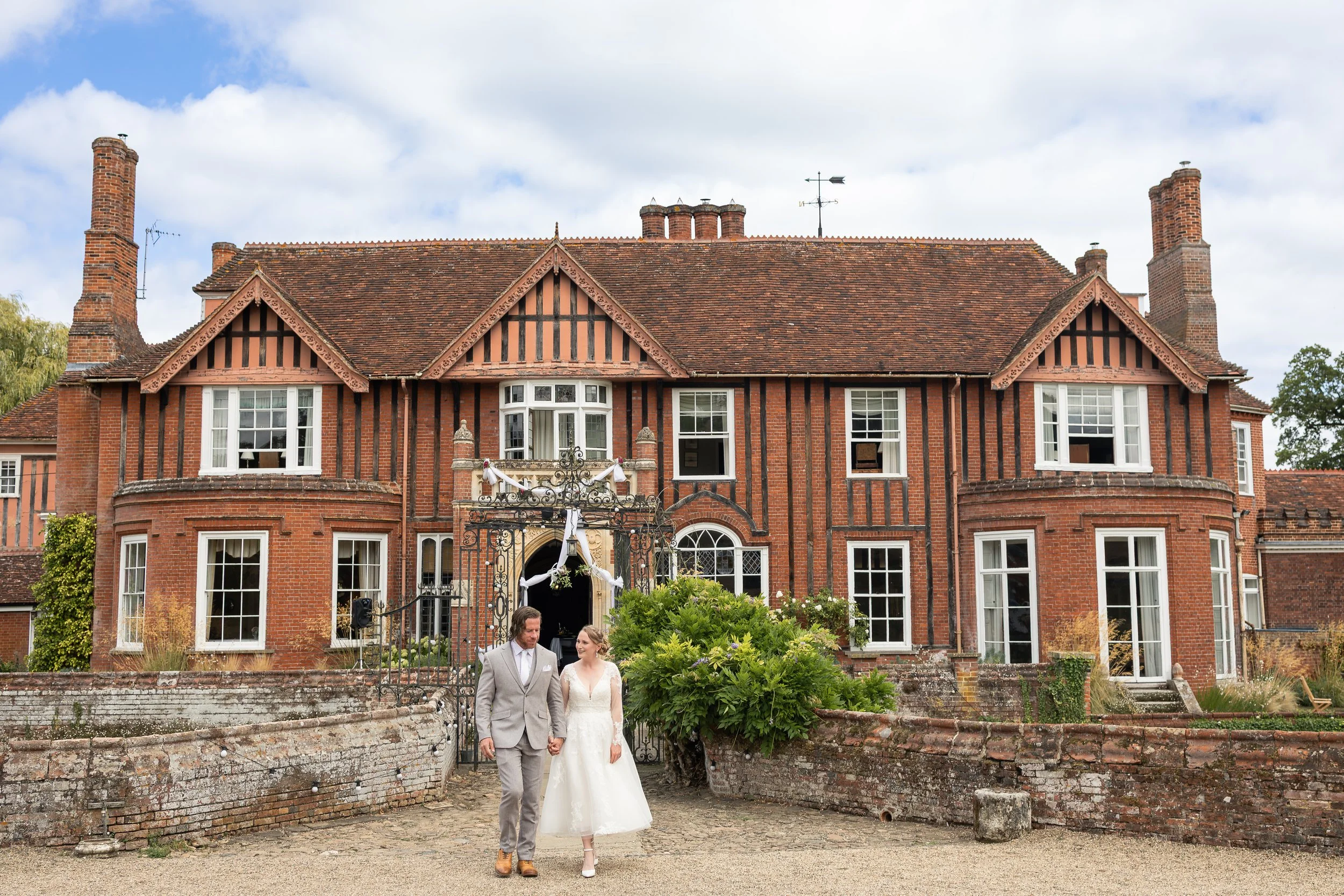A bride and groom walking hand in hand outside a large, historic brick house decorated for a wedding.