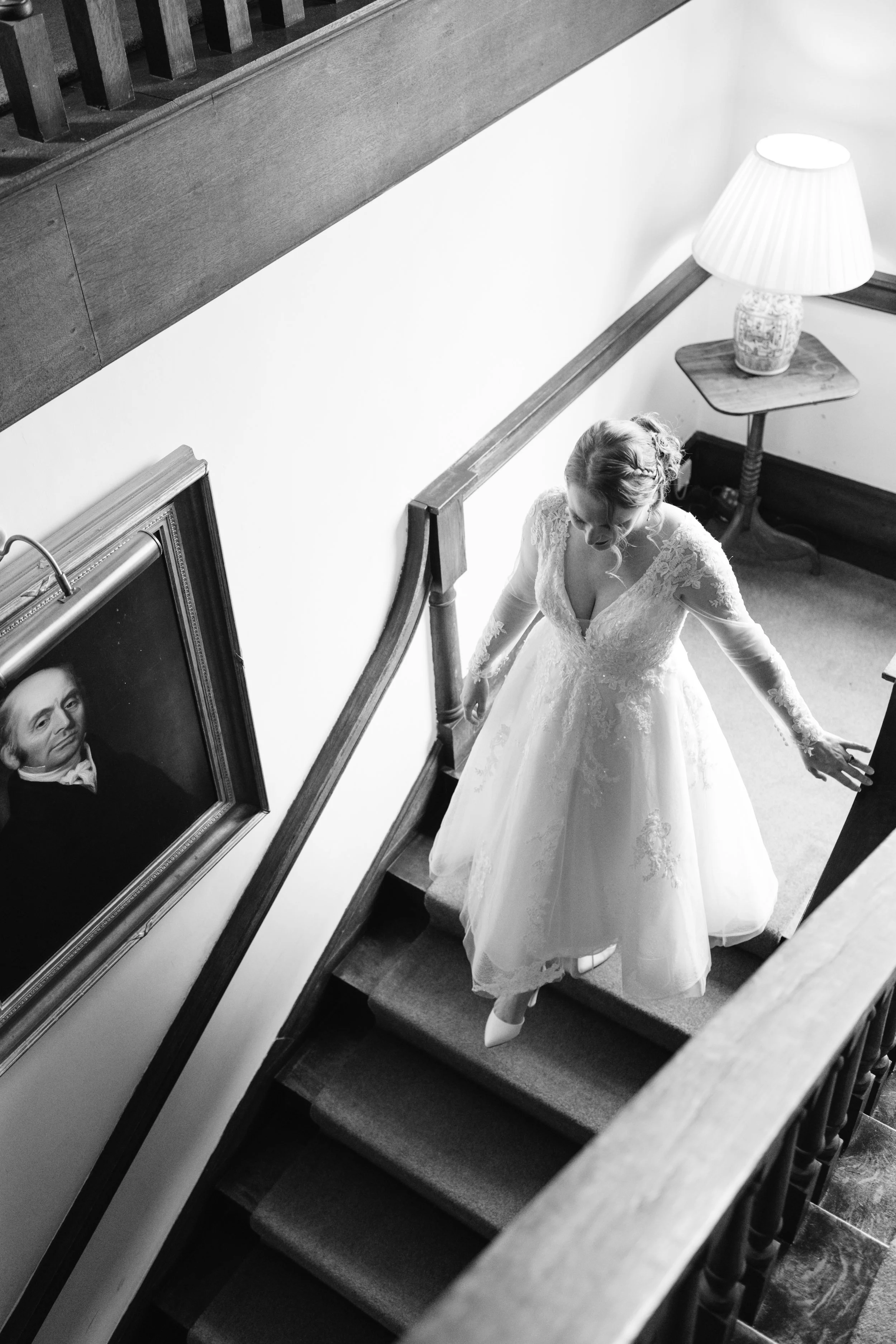 A bride descending a staircase in a wedding dress, holding the handrail with one hand, in a room with a framed portrait on the wall and a lamp on a small table.