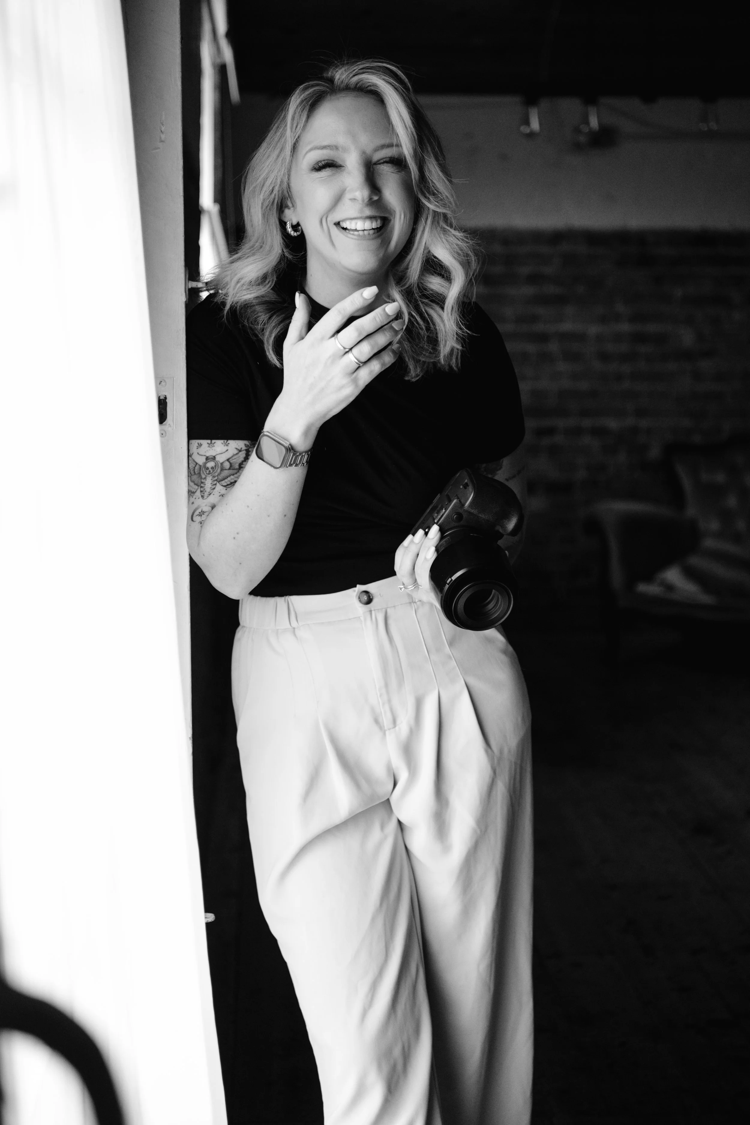 A woman with wavy hair, smiling and laughing, holding a camera and touching her face, wearing a black top, light-colored pants, jewelry, and a watch, standing indoors.