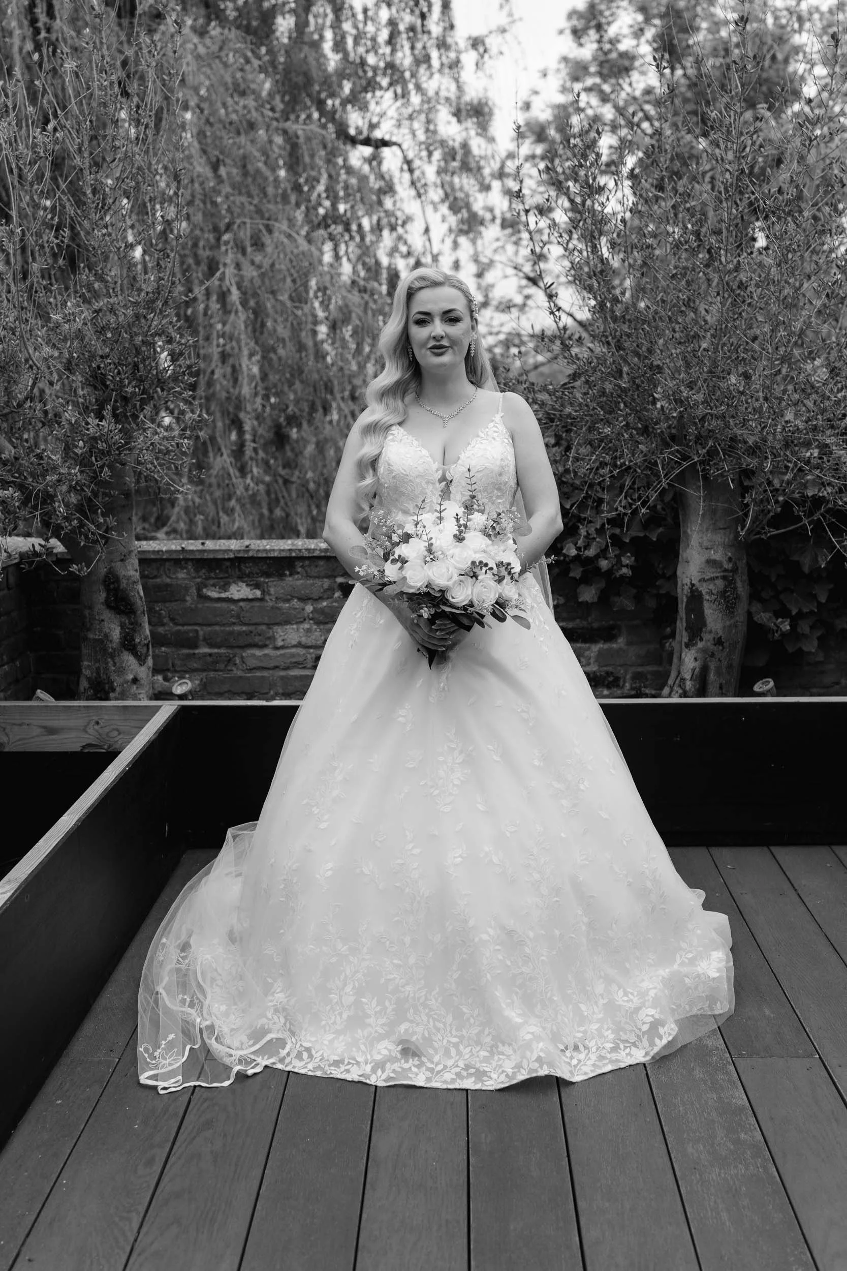 A bride standing outdoors on a wooden deck, holding a bouquet of flowers, wearing a wedding dress, with trees and foliage in the background.