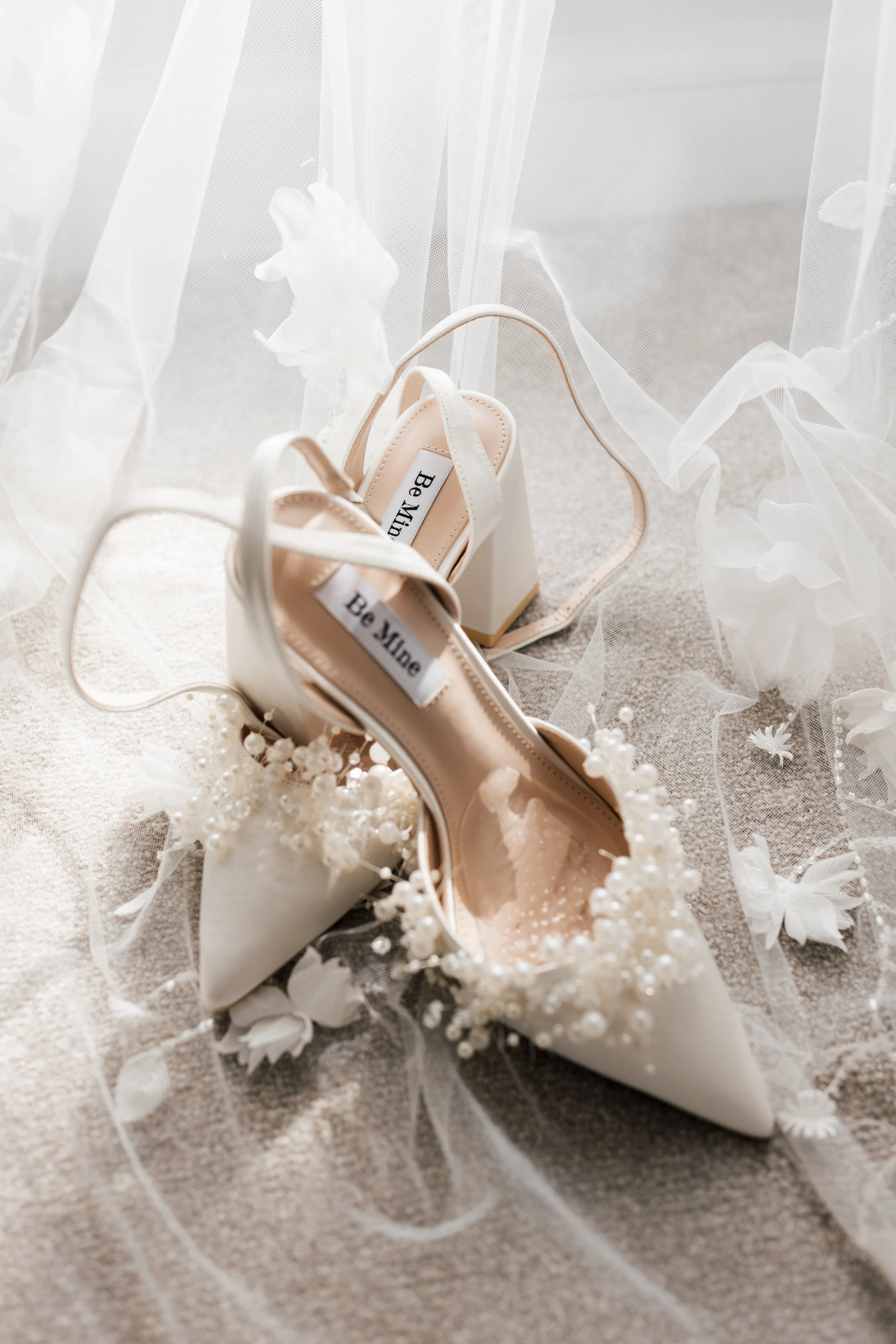A pair of white high-heeled shoes with the brand label 'Be Mine' inside, placed on a beige fabric surface, surrounded by delicate white floral decorations and translucent white fabric in the background.