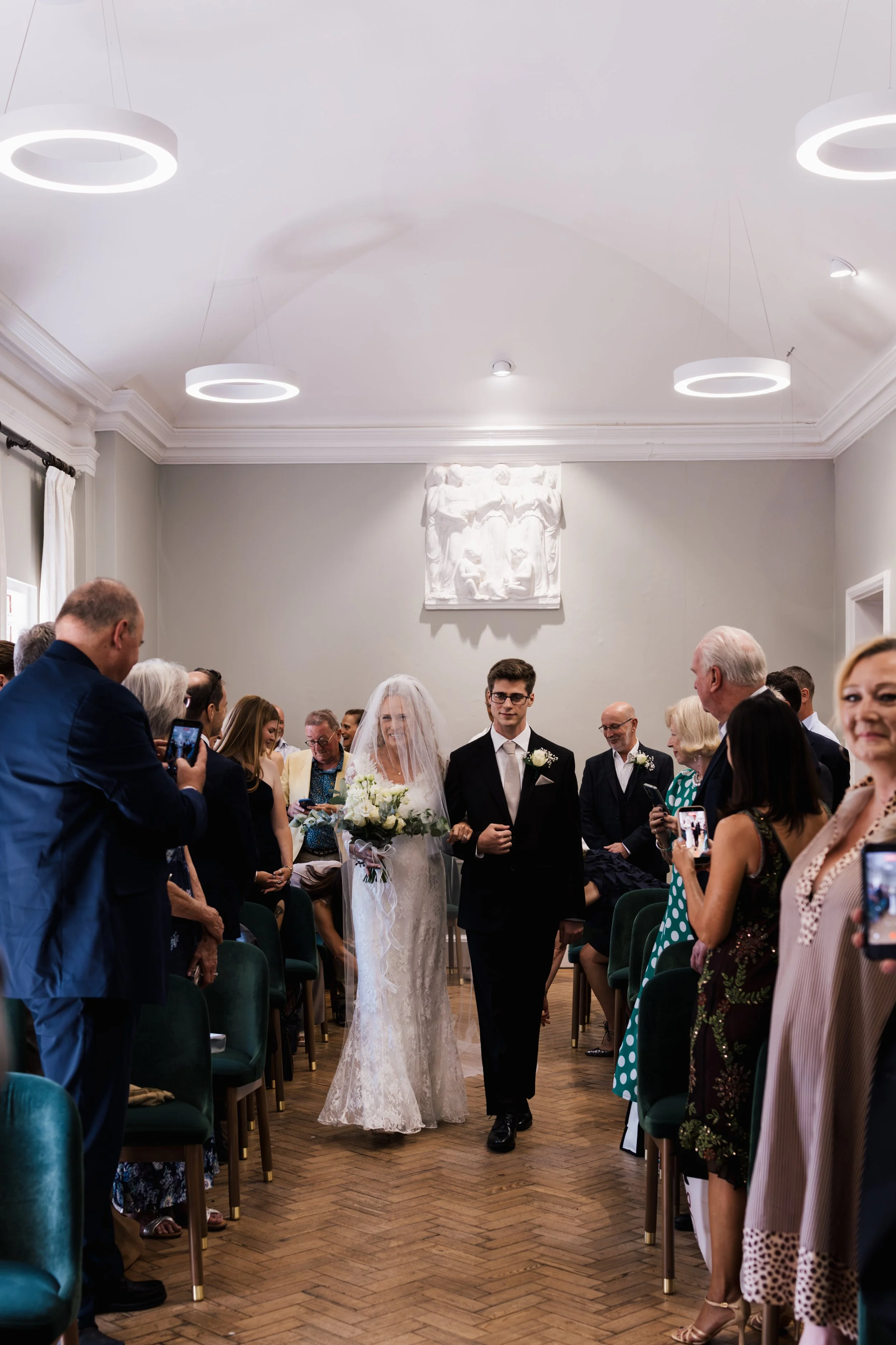 A bride and groom walking down the aisle in a wedding ceremony, surrounded by seated guests taking photos, with a white bas-relief sculpture on the wall behind them.