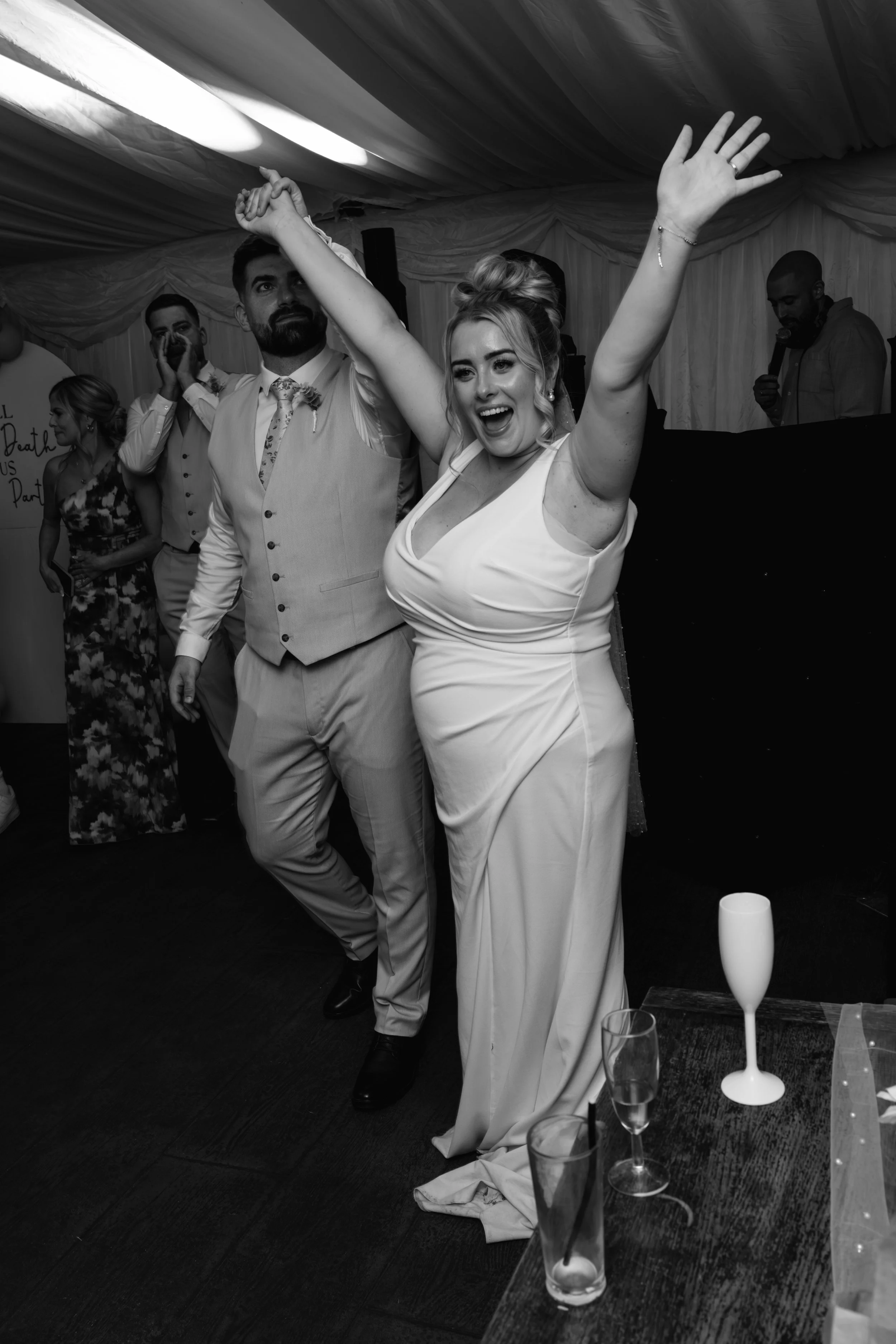 A woman with an updo hairstyle and a light-colored dress is dancing and smiling with her arms raised. Behind her, a man with a beard in a light suit and floral tie is also dancing. Other guests are visible in the background, some watching and others 