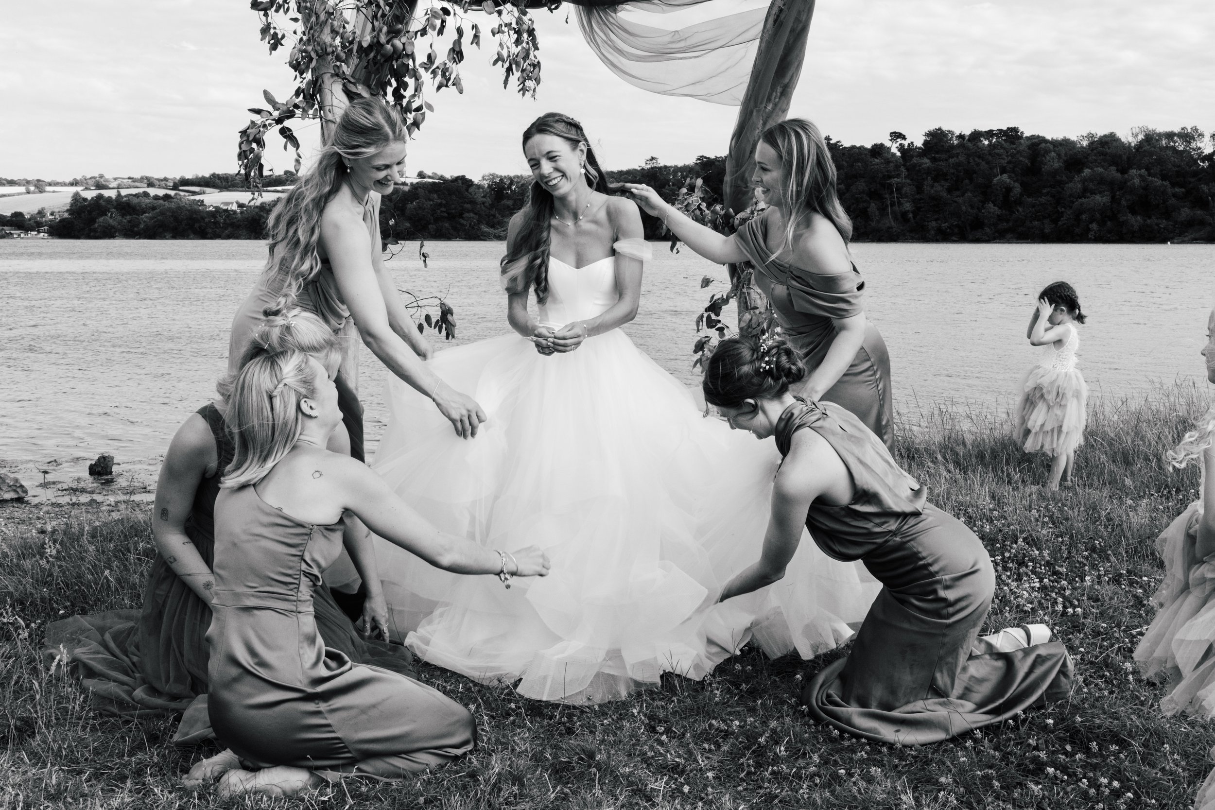 Black and white photo of a wedding party outdoors by a lake. The bride wears a strapless ball gown and is in the center, laughing. Surrounded by bridesmaids in matching dresses holding up the train of her dress, with some kneeling and some standing. 