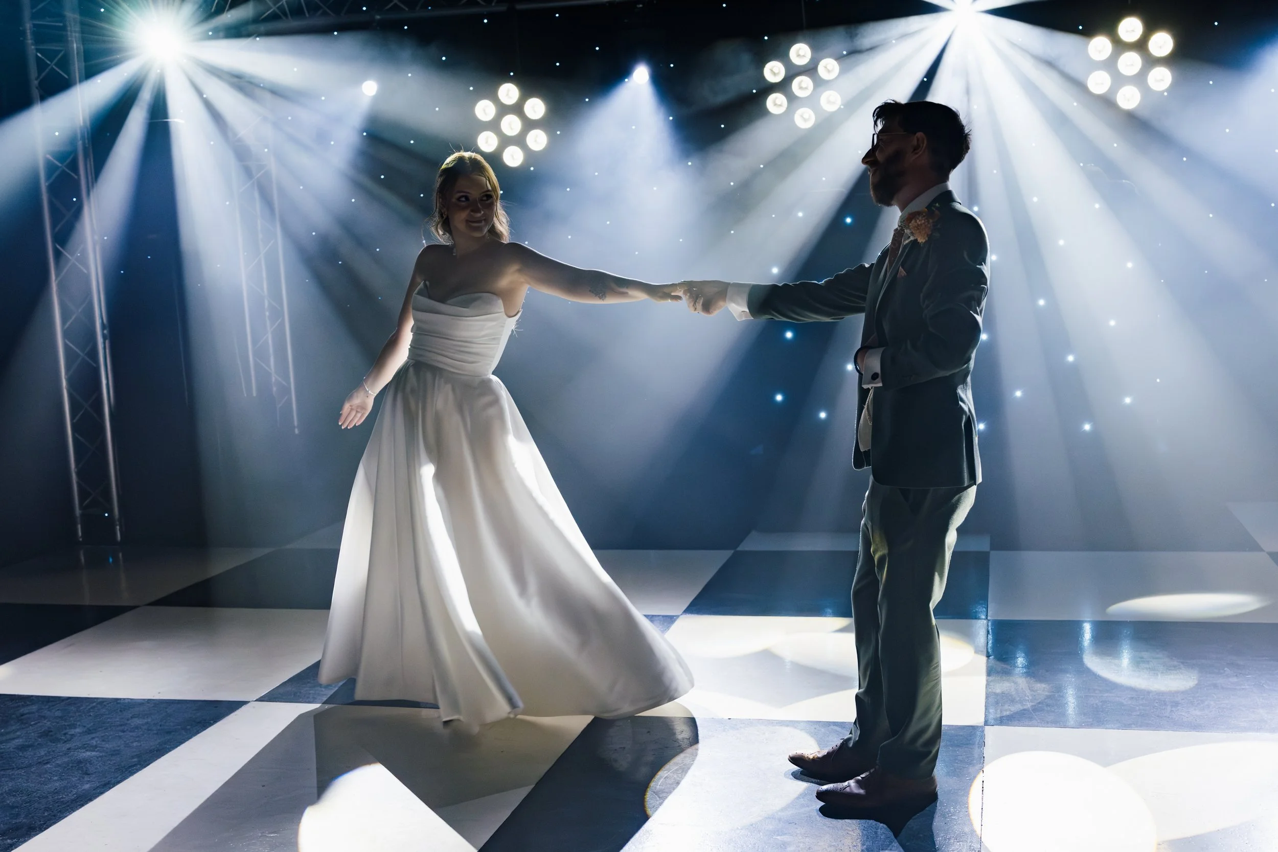 A bride and groom dancing at their wedding reception, holding hands on a checkered dance floor with dramatic lighting and spotlights overhead.
