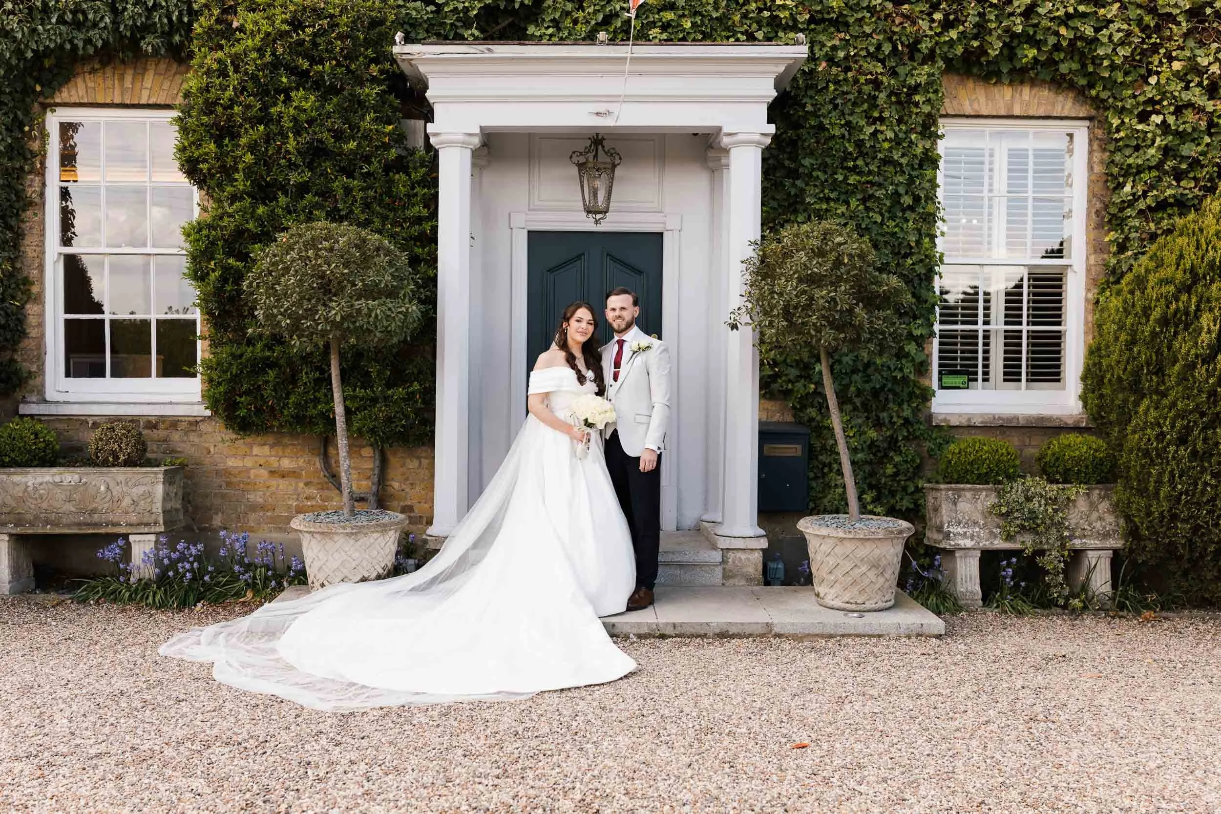 A bride and groom standing together in front of a house with ivy-covered walls, large windows, and potted plants. The bride is wearing a white wedding dress with a long train and holding a bouquet, and the groom is wearing a white suit with a red tie