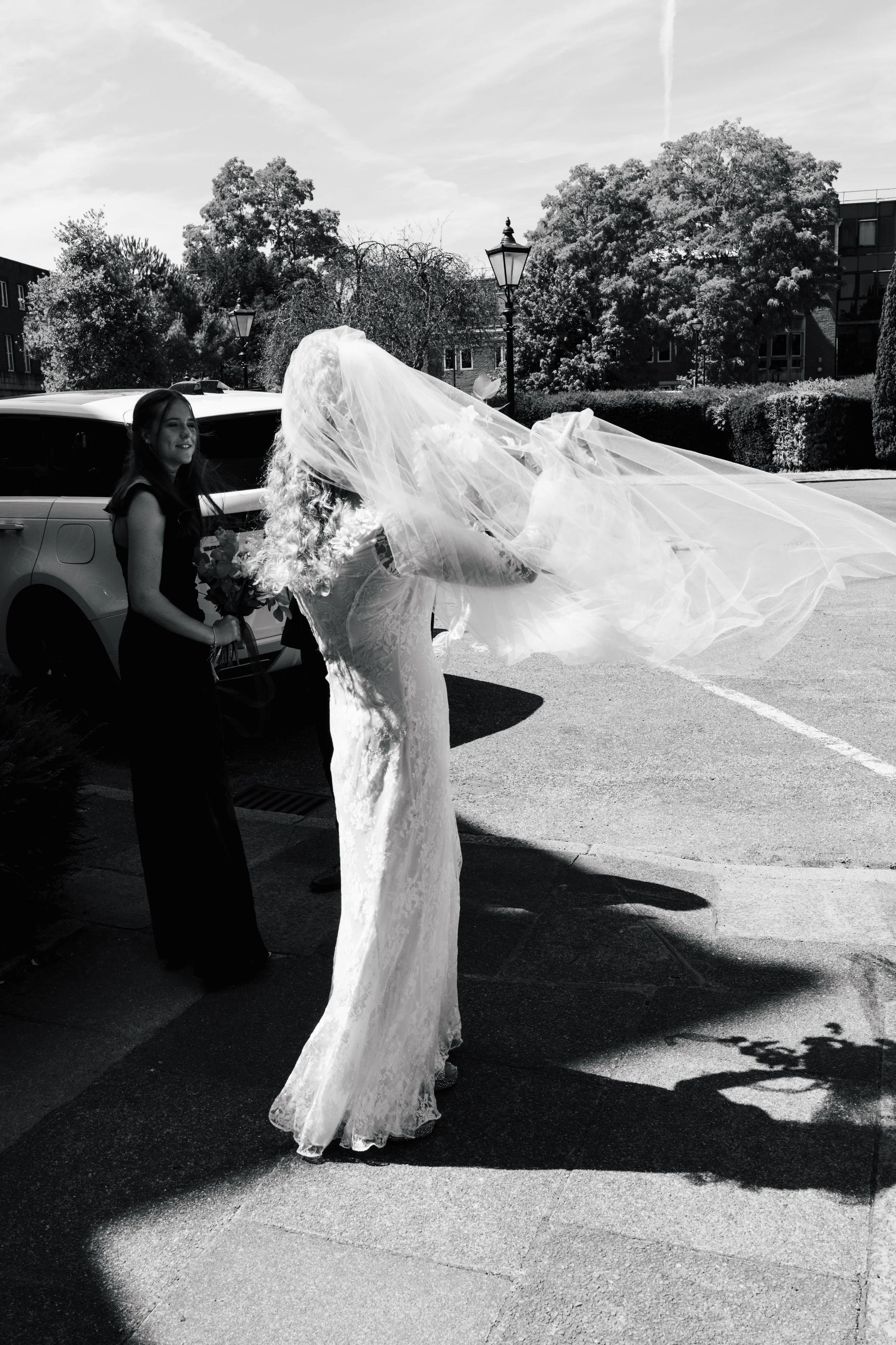A bride in a wedding dress and veil standing outside, holding a bouquet, with a young girl in a dark dress beside her, near a car, trees, and lamp posts on a sunny day.