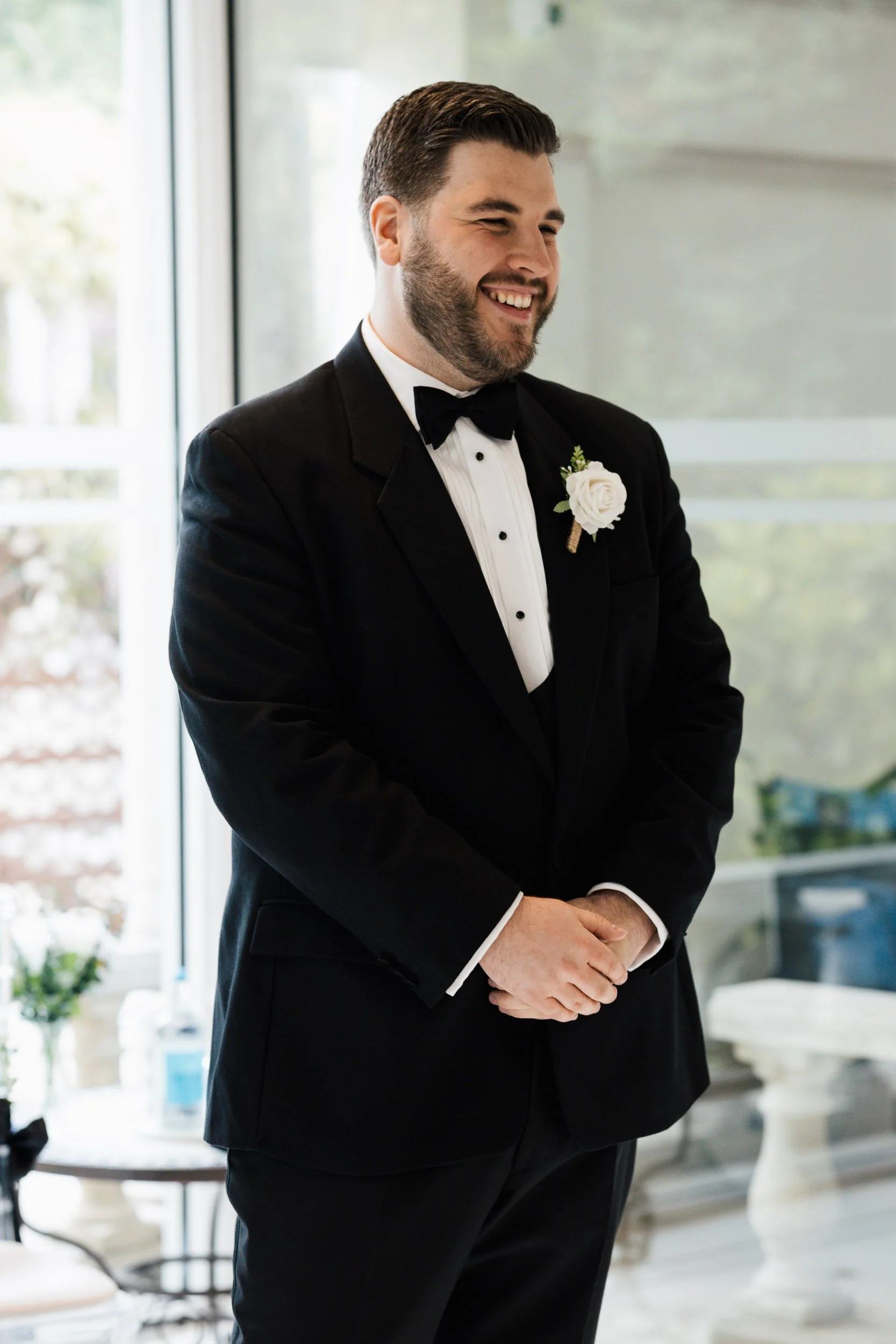 A man dressed in a tuxedo with a white shirt, black bow tie, and a white boutonniere, standing indoors with a big smile, possibly at a wedding or formal event.
