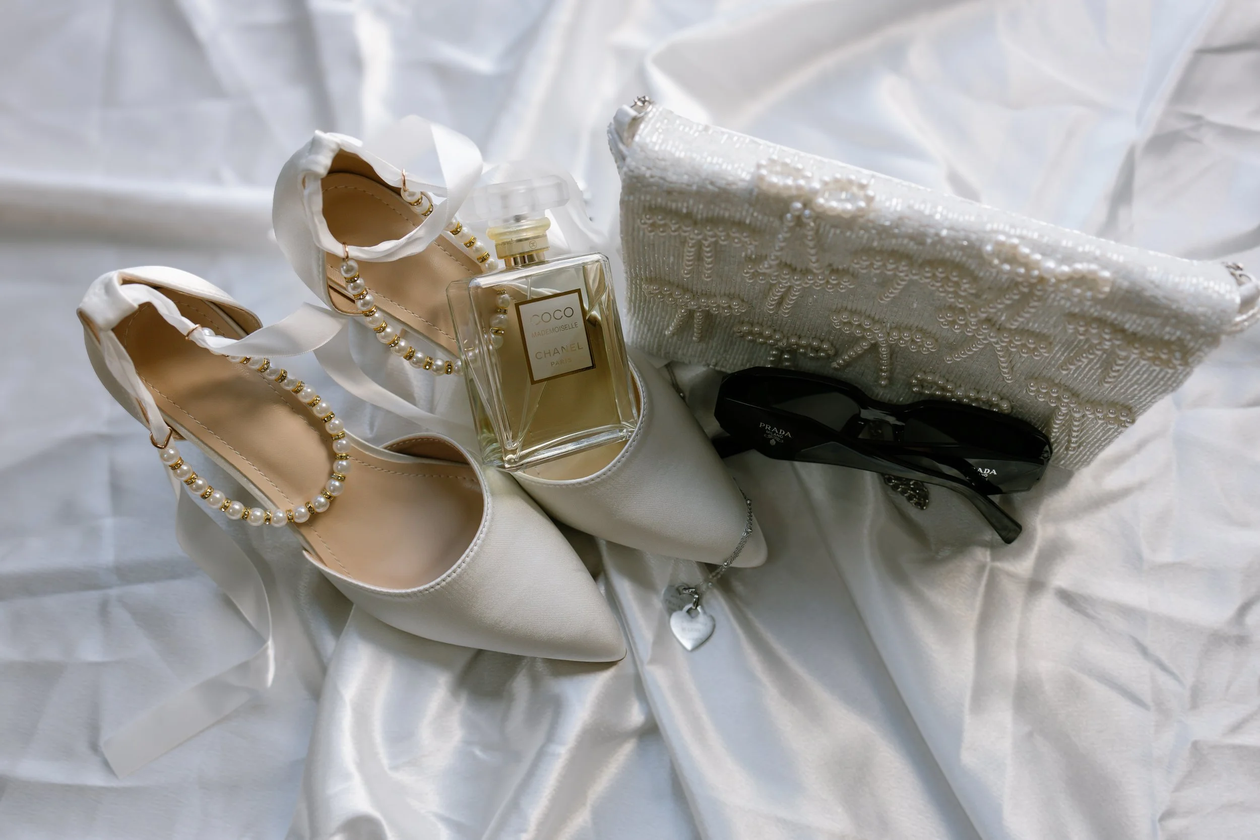 White high-heeled shoes with pearl and gold embellishments, a bottle of Chanel No. 5 perfume, a pearl-embellished clutch, black Prada sunglasses, and a silver heart necklace, all on white satin fabric.