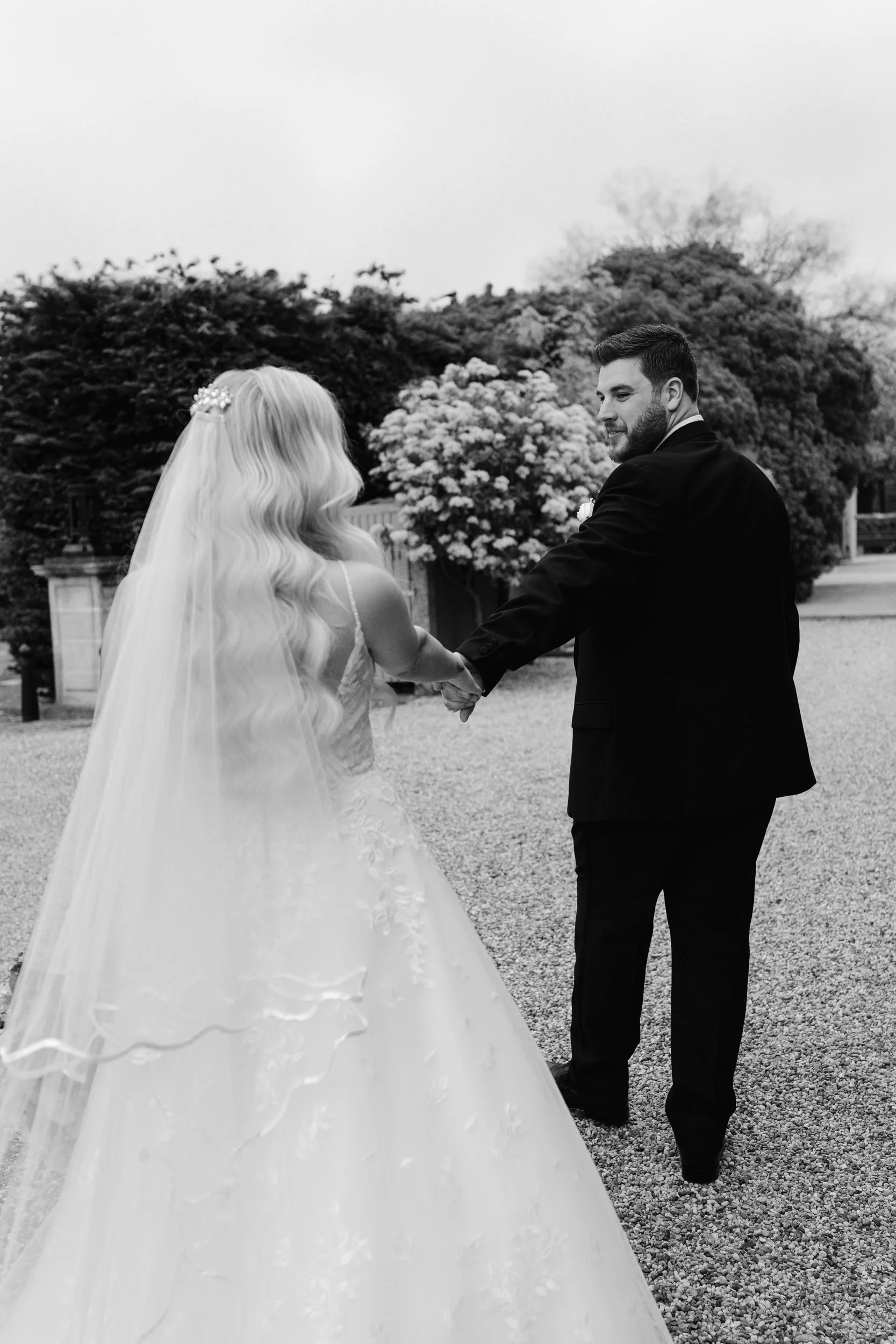 Black and white photo of a bride and groom holding hands outside. The bride has long wavy hair and a veil, the groom has short dark hair and a beard, wearing a suit. There are bushes, trees, and a cloudy sky in the background.