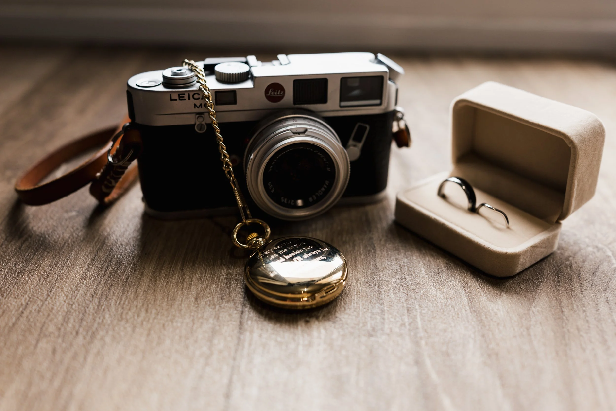 A vintage film camera with a brown leather strap, a gold chain, and a watch in a beige jewelry case on a wooden surface.