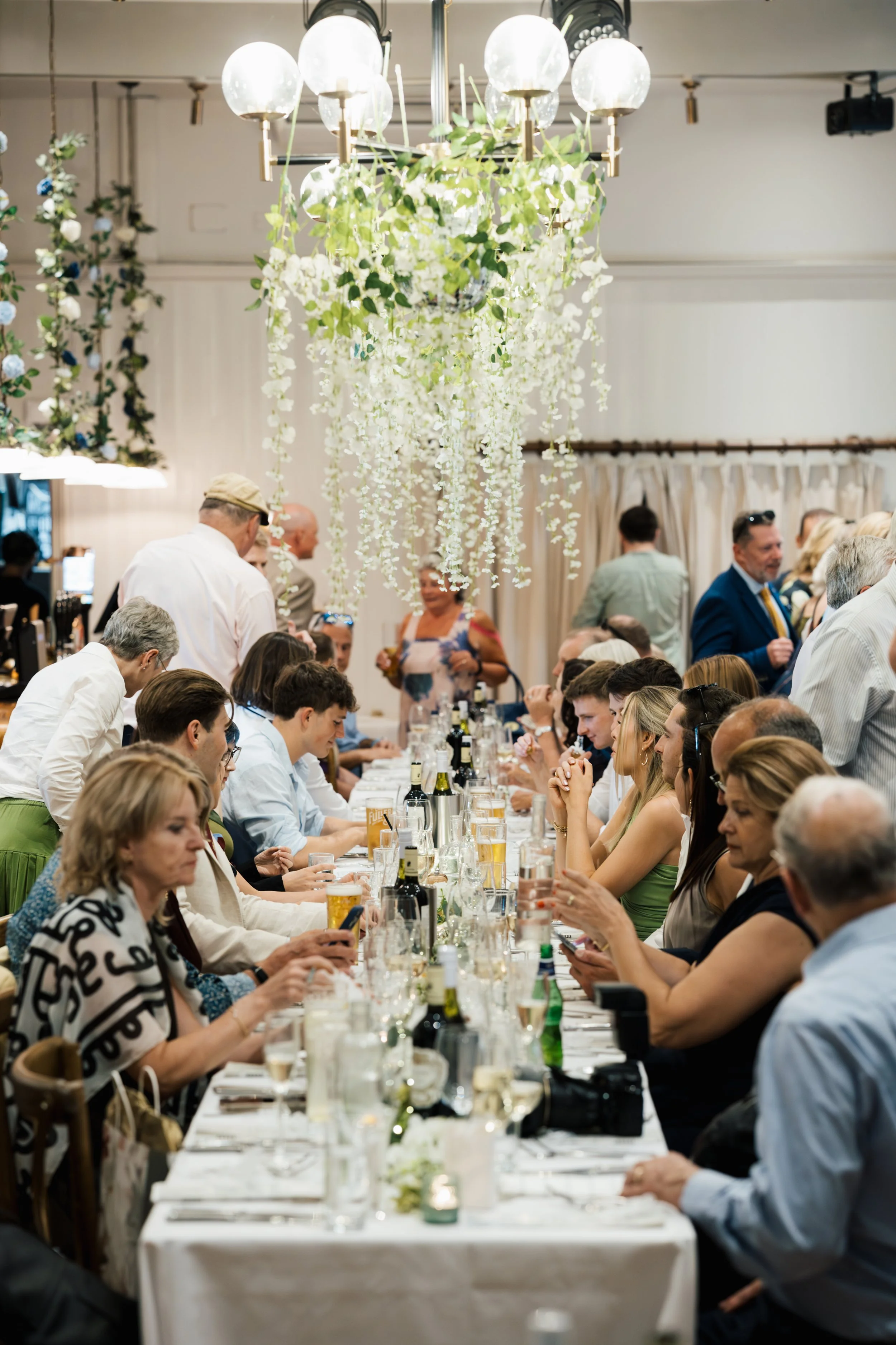 People gathered around a long dining table at a formal event, with white tablecloth, wine bottles, and glasses. Chandeliers with hanging flowers are overhead, and some attendees are talking, using their phones, or eating.