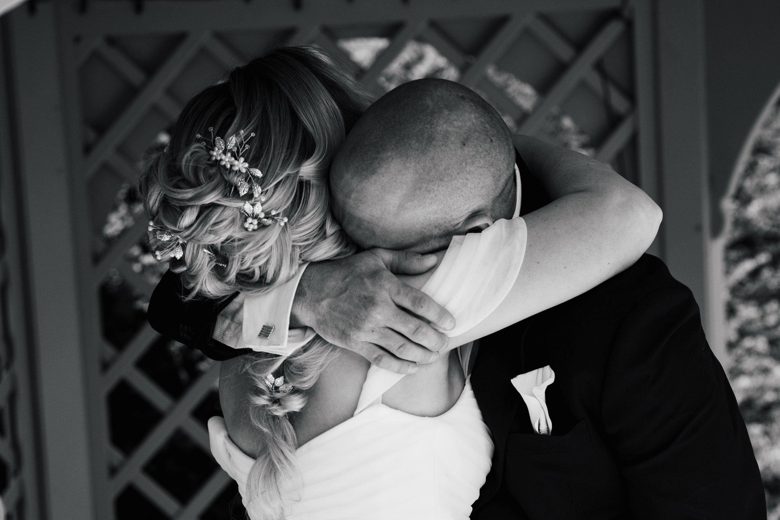 A woman and a man sharing an embrace, with the woman wearing a floral hair accessory and the man having a shaved head, both in formal attire, outdoors under a wooden lattice structure.